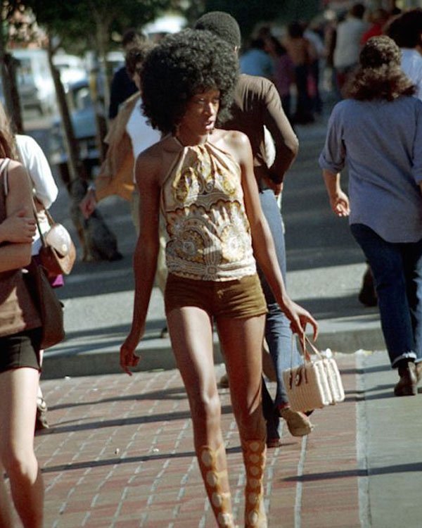 Pessoa andando com roupa e cabelo característicos dos anos 70, destacando um top estampado e botas de plataforma.