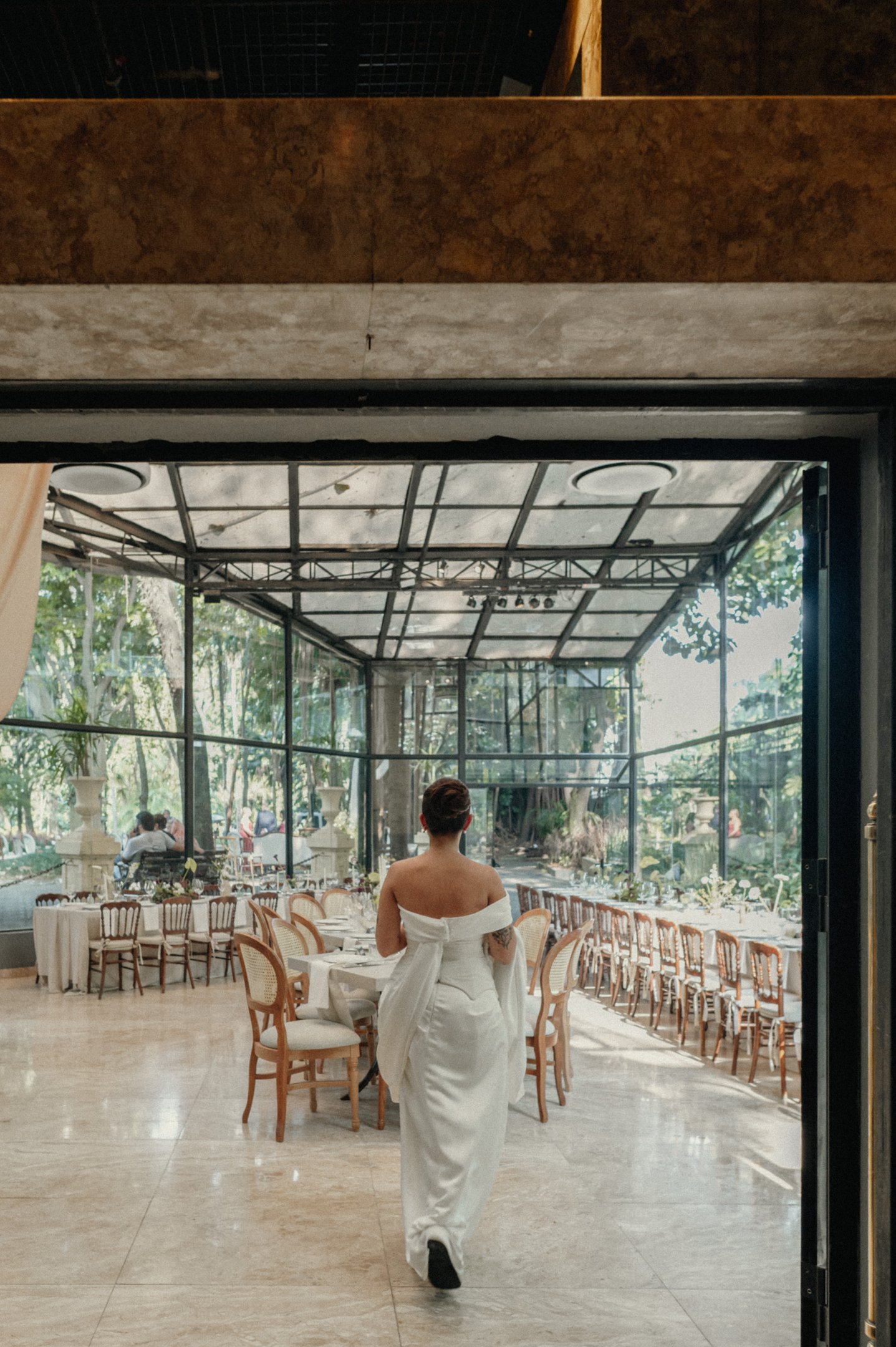 Mulher usando vestido branco com cauda em ambiente de festa elegante. Visual sofisticado com ombros à mostra e cabelo preso. Tendência clássica e atemporal para eventos formais, destacando uma estética minimalista e refinada.