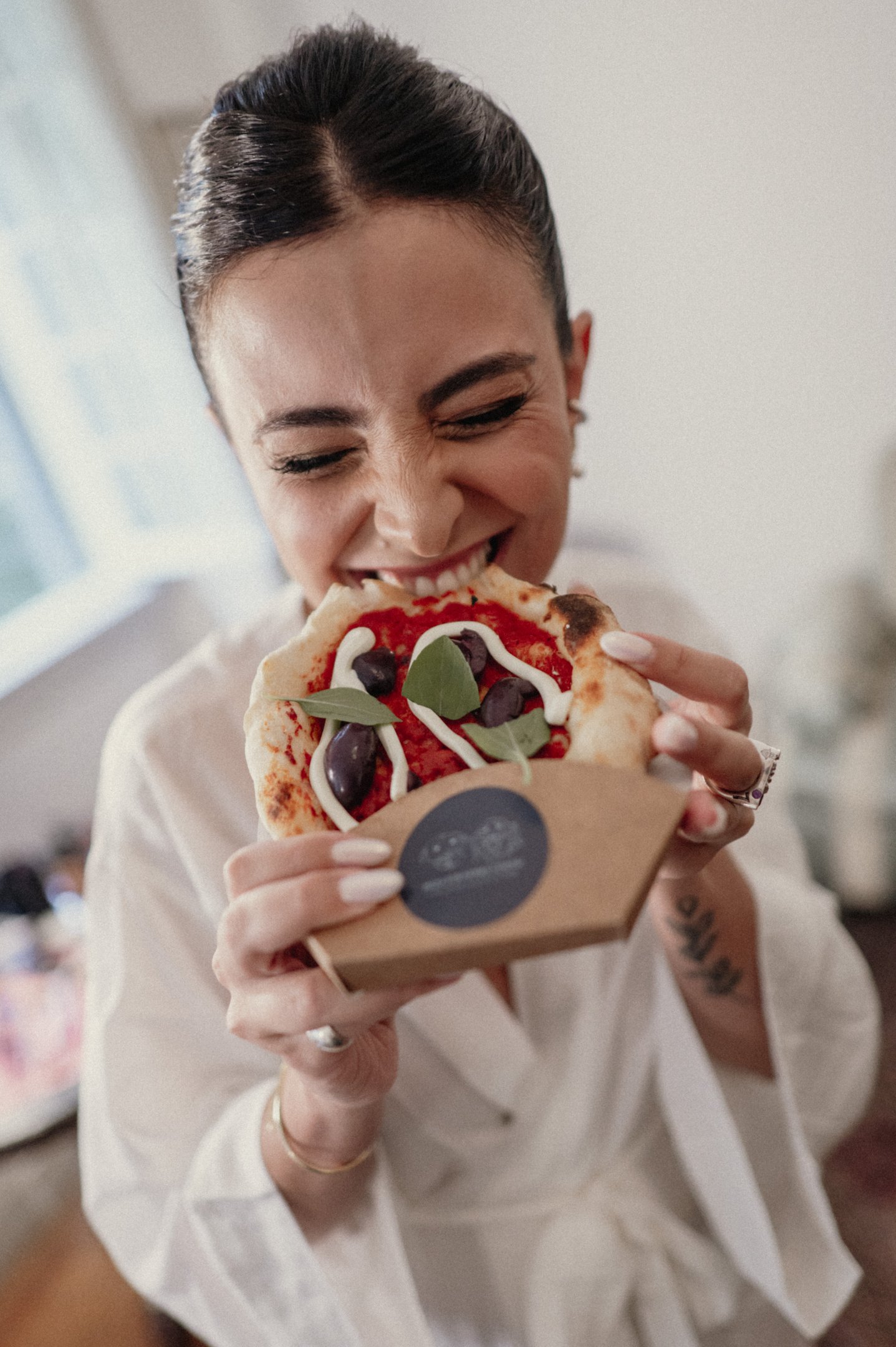 Lela Brandão com cabelo preso e unhas bem cuidadas veste um robe branco, tendência minimalista e elegante. Ela segura uma pequena pizza, destacando um estilo descontraído e moderno, combinando conforto e sofisticação.