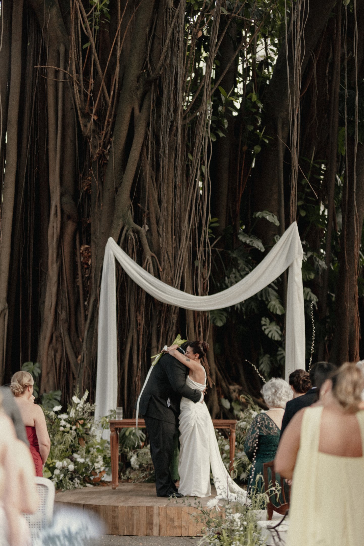 Lela Brandão e Viktor Murer em cerimônia ao ar livre com decoração minimalista. A noiva veste um vestido branco clássico e o noivo, terno preto tradicional. Convidados em trajes formais, com destaque para vestidos em tons neutros e cortes elegantes.