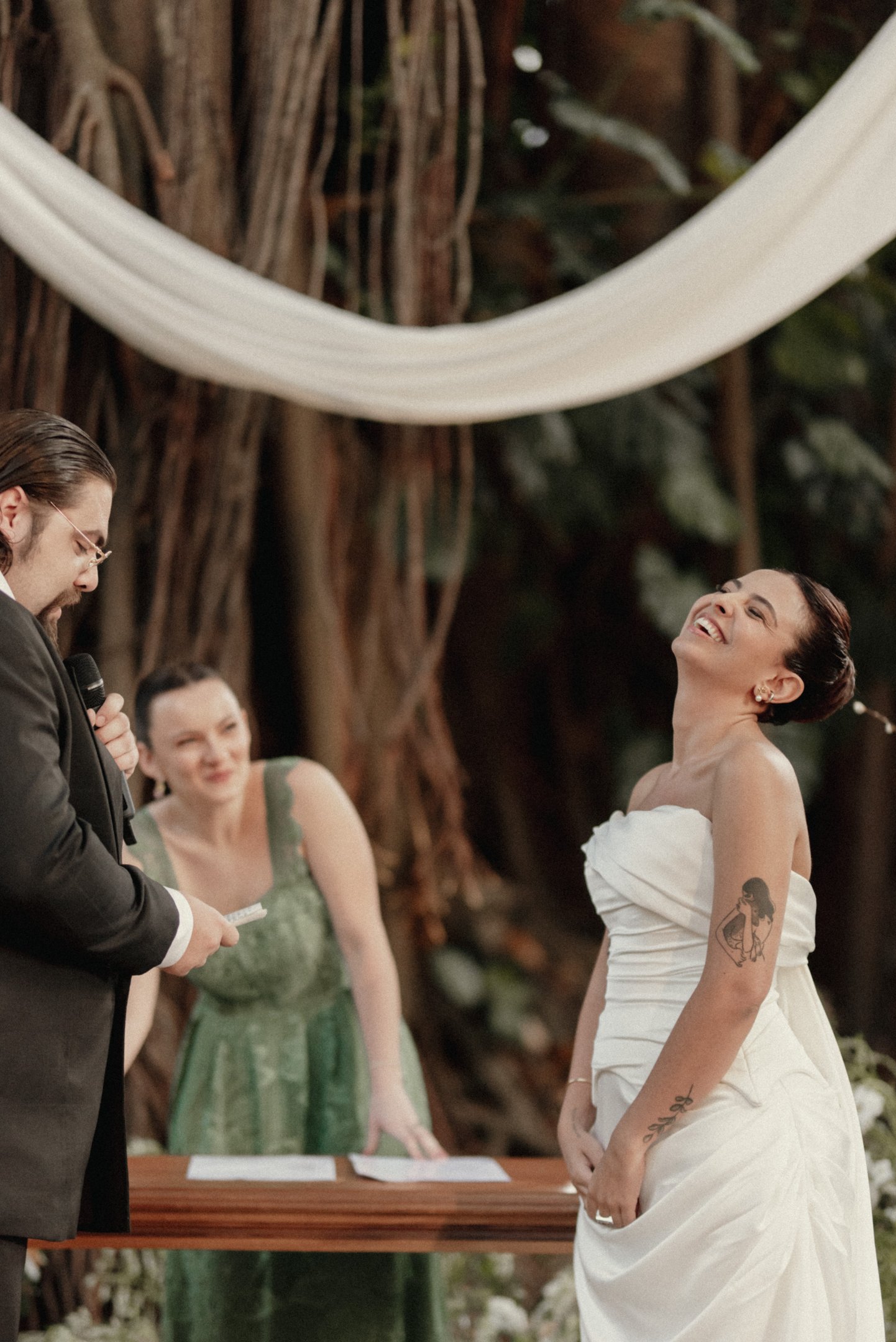 Casamento ao ar livre com noiva em vestido branco clássico, drapeado sem alças. Convidada usa vestido verde rendado, estilo romântico. Noivo veste terno preto elegante. Estilo descontraído e sofisticado em cerimônia rústica.