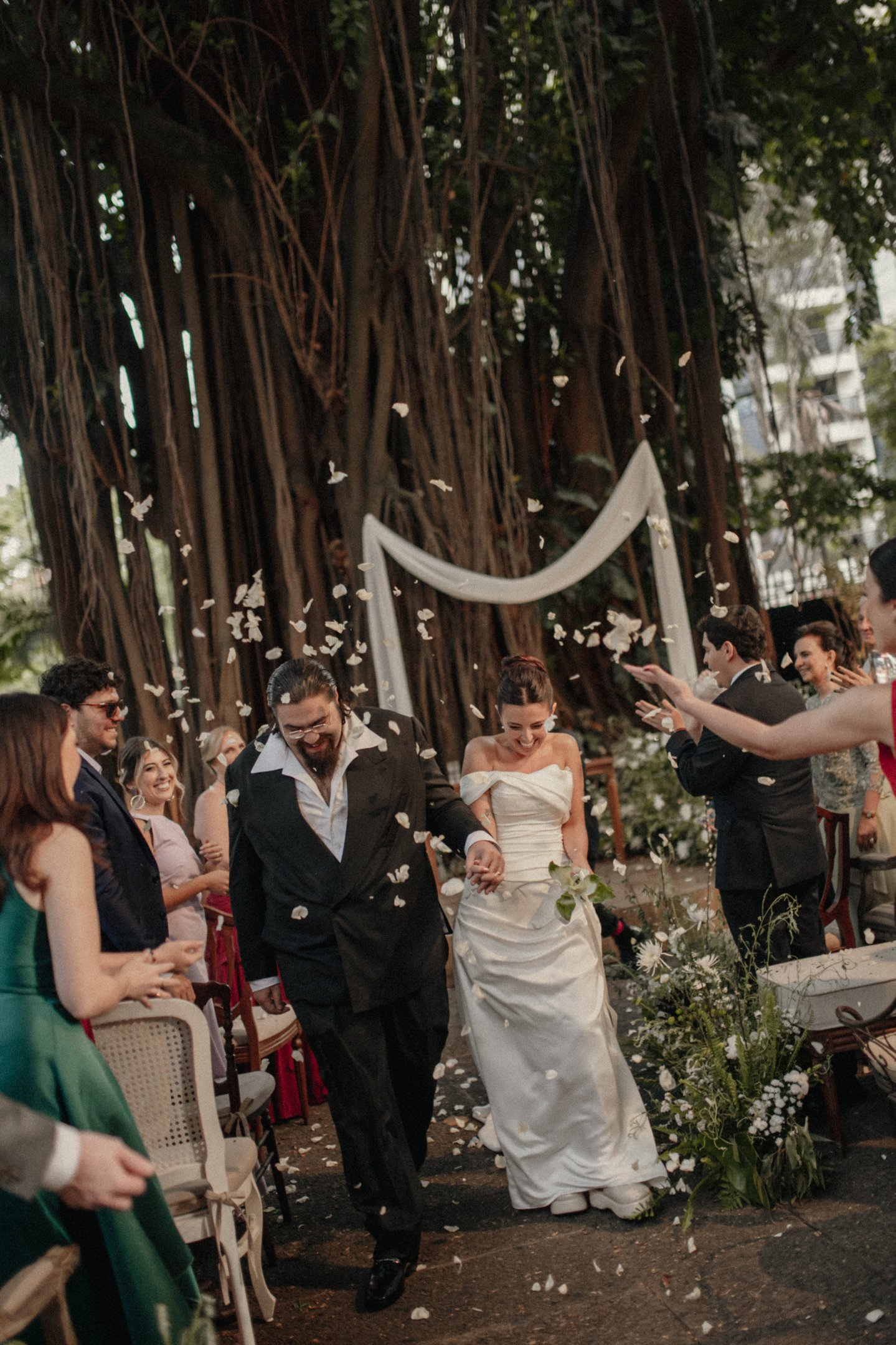 Lela Brandão em casamento ao ar livre. Noiva com vestido tomara que caia e cabelo preso em coque elegante. Noivo de terno preto clássico e camisa branca. Convidados em roupas de festa sofisticadas, cores vivas e tecidos leves.