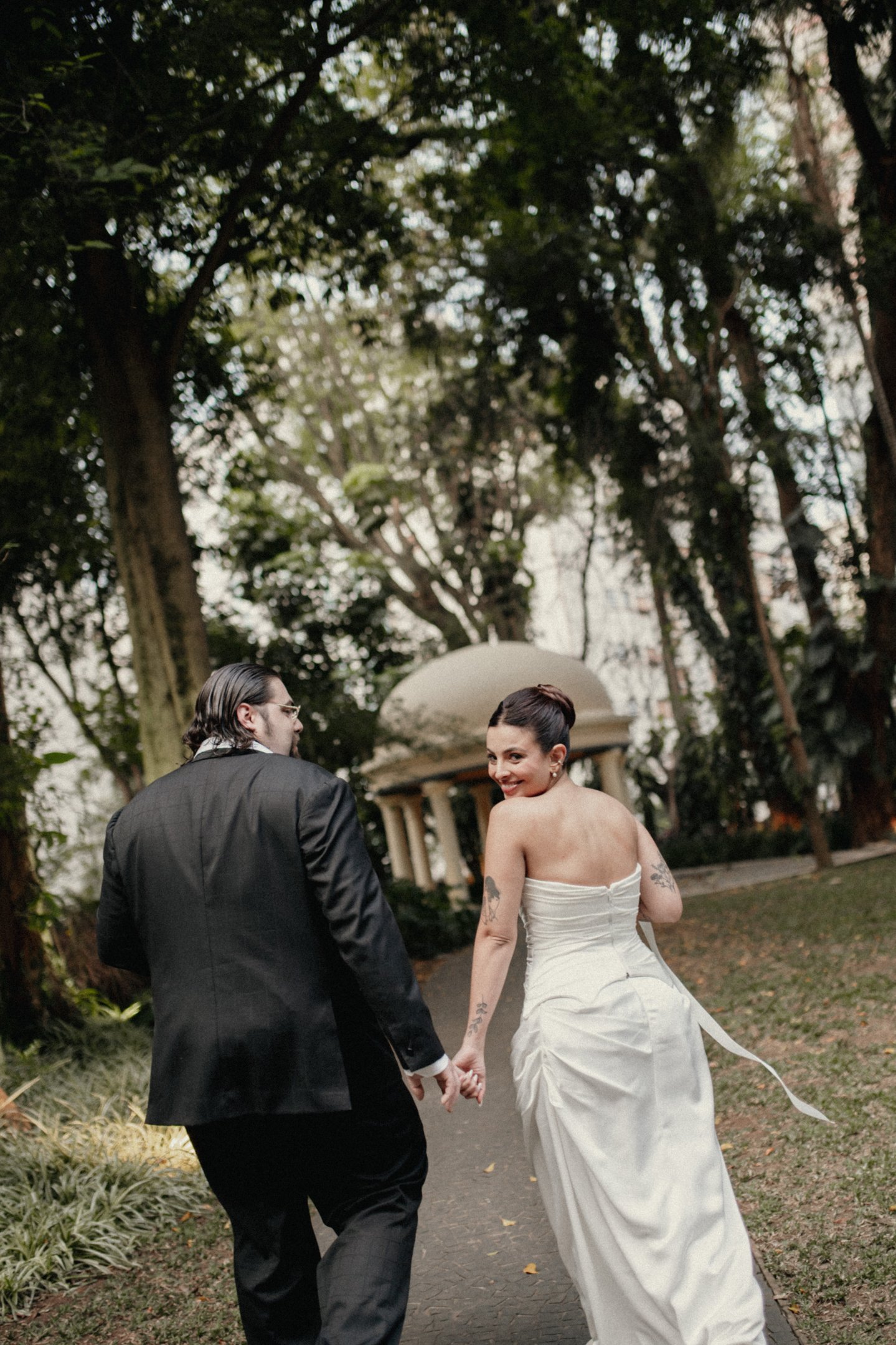 Casal em traje de casamento, com o noivo vestindo um elegante terno preto e a noiva em um vestido branco estilo clássico, sem alças. A composição destaca um ambiente natural, tendência de casamentos ao ar livre e elegância atemporal.