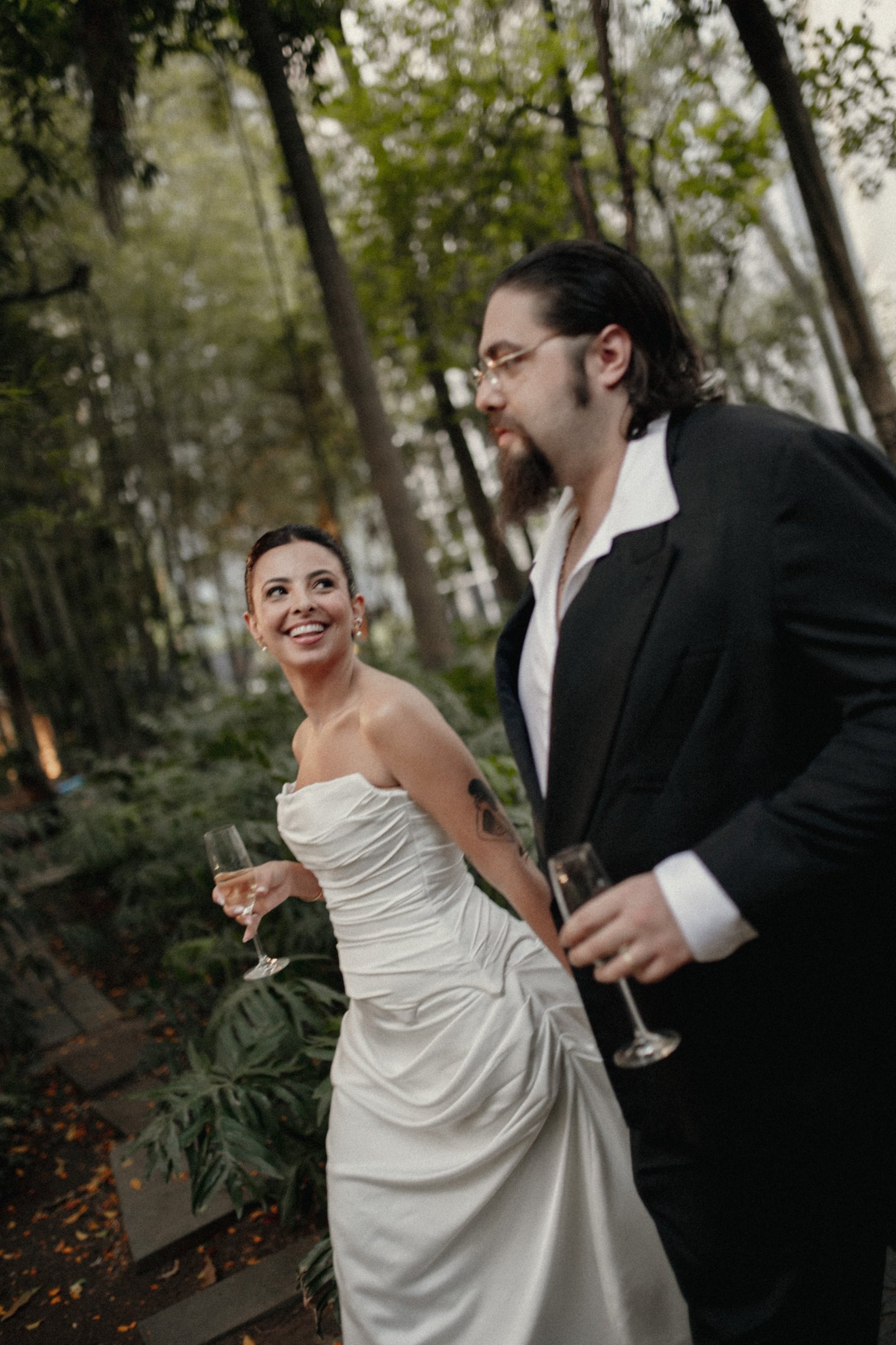 Casal em traje nupcial moderno, vestido de noiva com drapeados e cauda elegante; noivo em terno preto clássico com camisa branca aberta. Estilo sofisticado, combinando elementos tradicionais e contemporâneos, ideal para casamentos ao ar livre.