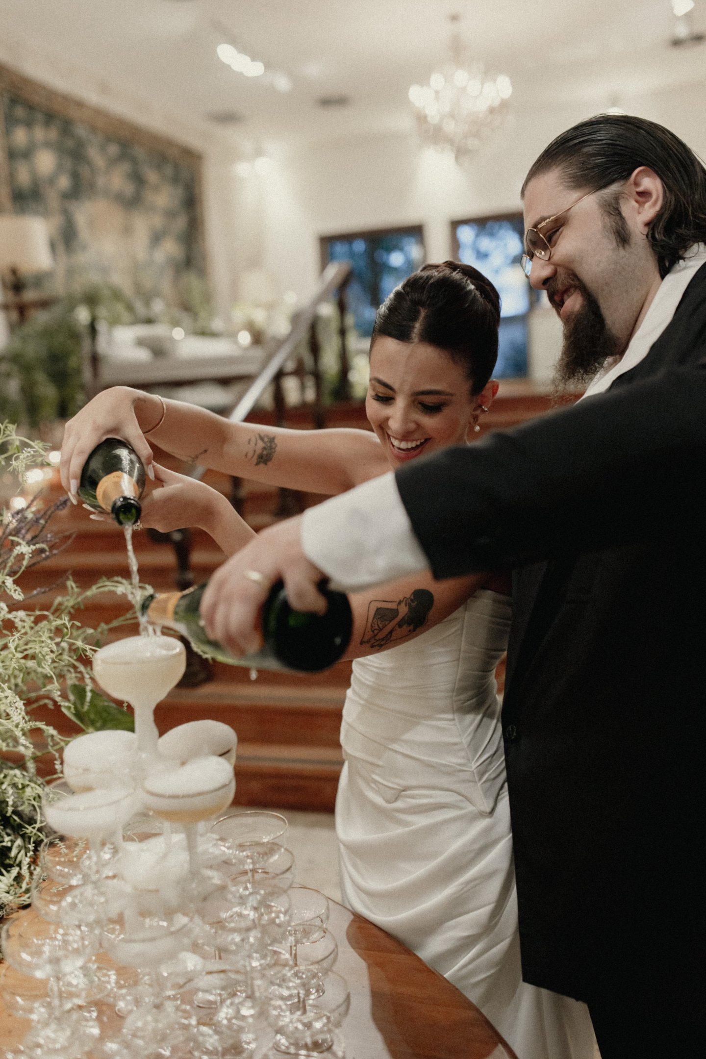 Casal elegante celebra com champanhe. Ela veste vestido de noiva clássico com detalhes drapeados, e penteado sofisticado. Ele, em terno preto e camisa branca, exibe estilo tradicional. Ambiente sugere evento formal e festivo.