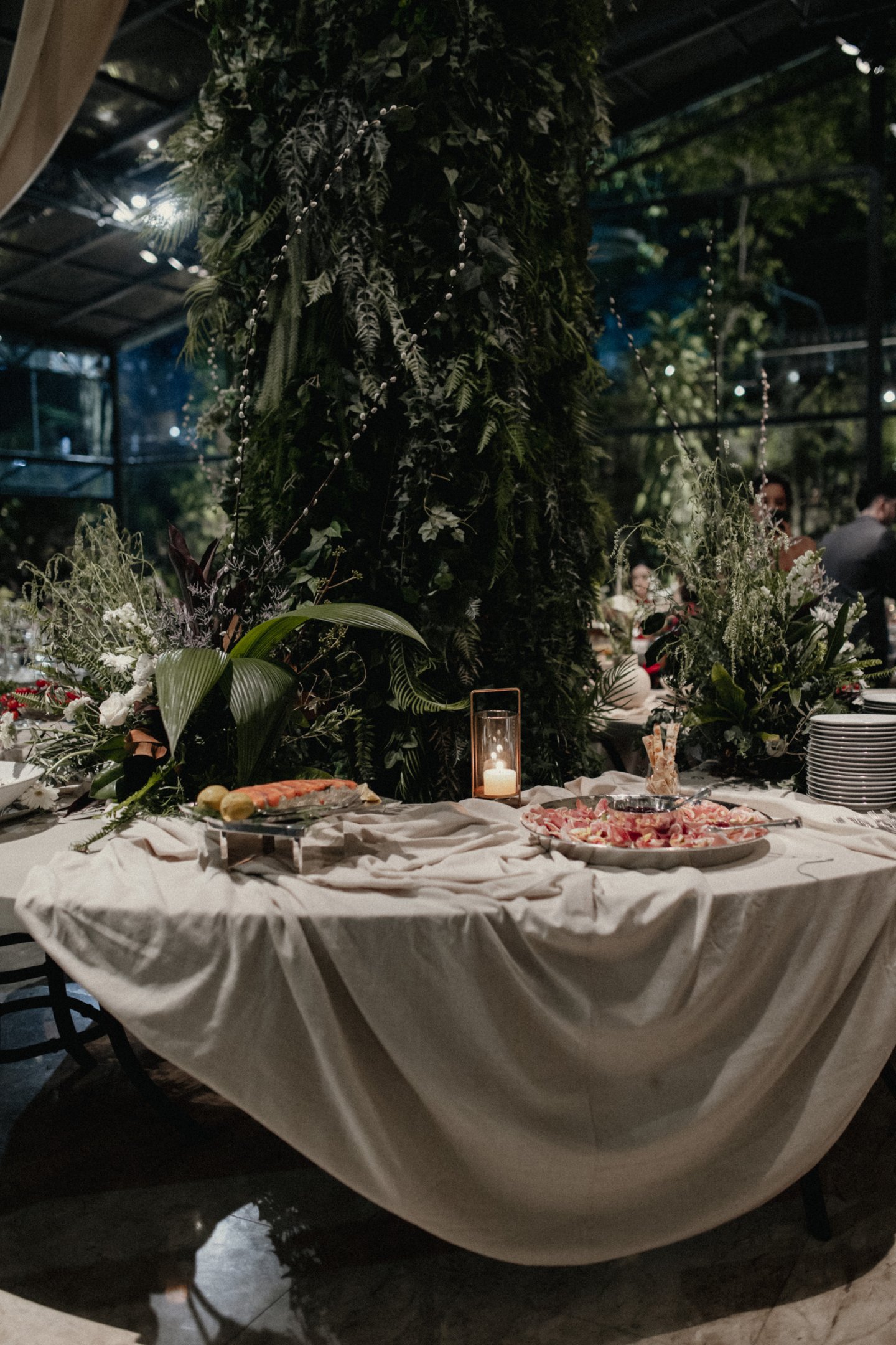 Mesa de buffet sofisticada com toalhas de linho drapeadas, rodeada por decoração verde exuberante. Ambiente elegante e natural, ideal para eventos ao ar livre com tendências de moda sustentáveis e minimalistas.