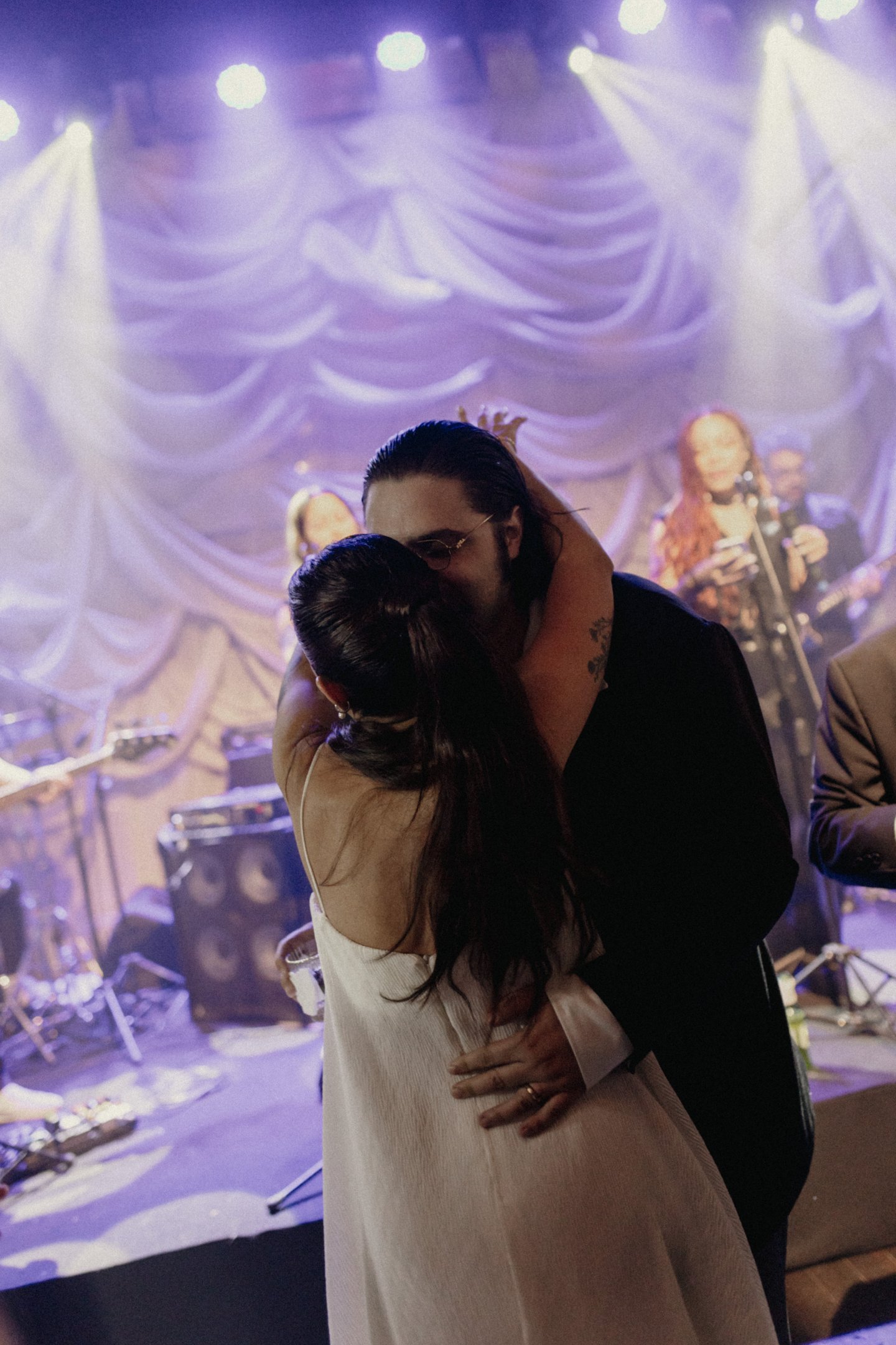 Casal elegante dançando em evento. Mulher veste vestido branco minimalista e solto, enquanto o homem usa terno preto clássico. A atmosfera é de festa, com iluminação suave e músicos ao fundo. Tendência de moda sofisticada e atemporal.