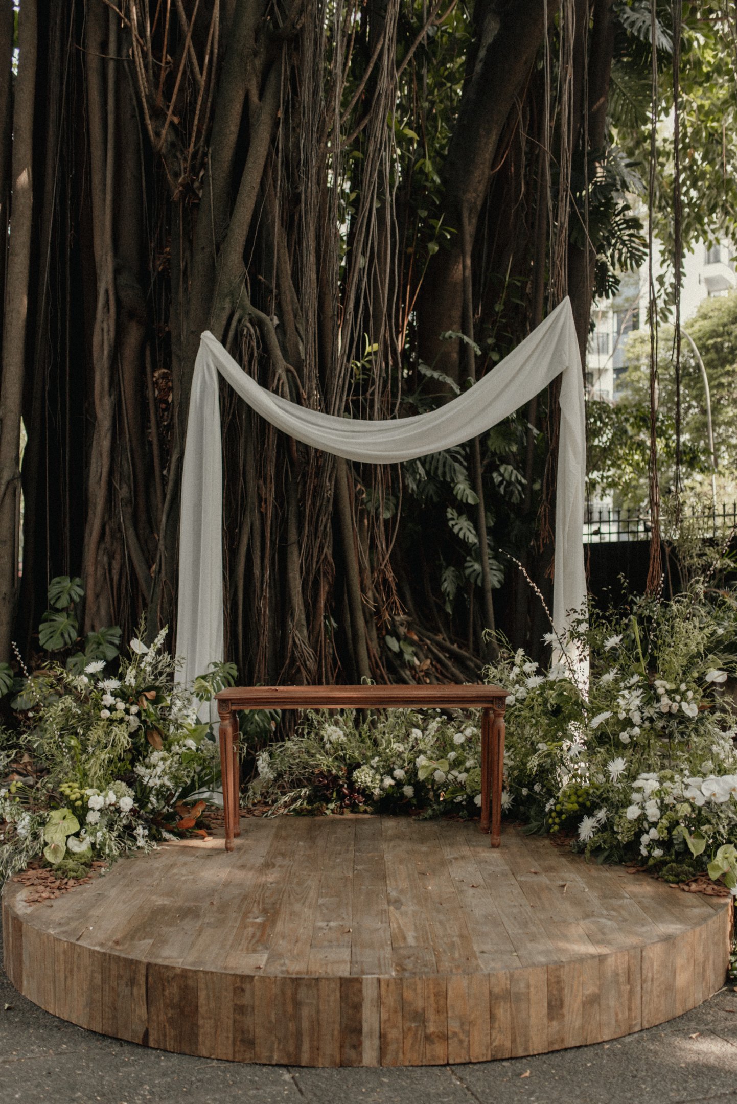 Espaço de cerimônia ao ar livre com decoração minimalista. Estrutura de madeira cercada por flores brancas e folhagens, complementada por tecido branco pendurado em árvores. Casamento Lela Brandão.