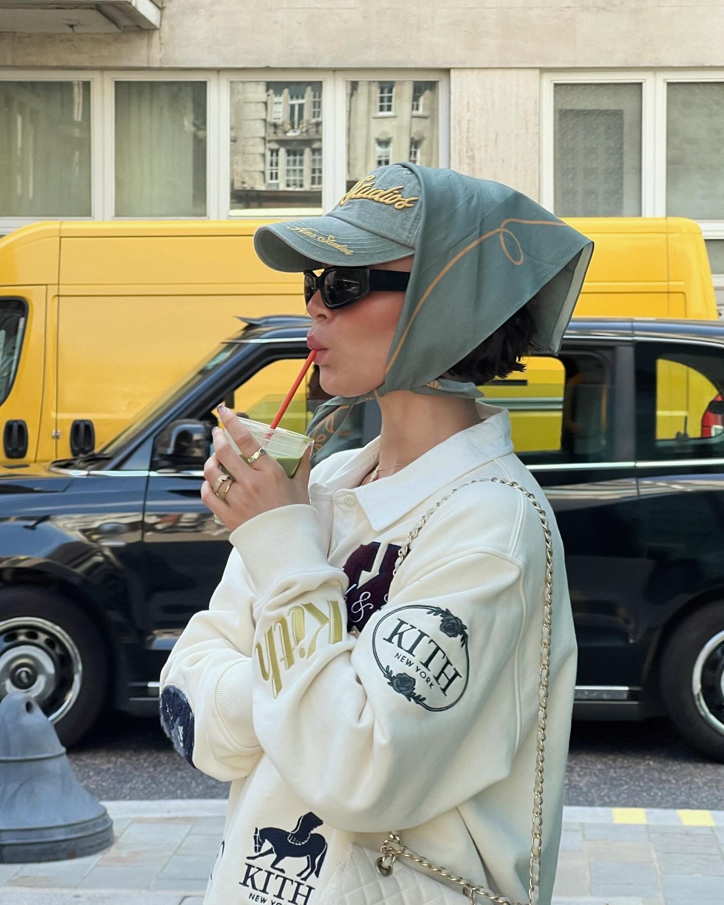 Pessoa vestindo moletom oversized com logotipo Kith, boné estilizado e lenço na cabeça. Óculos escuros completam o look urbano e moderno. Estilo casual-chic, misturando acessórios esportivos e retrô. Trendy e descontraído.