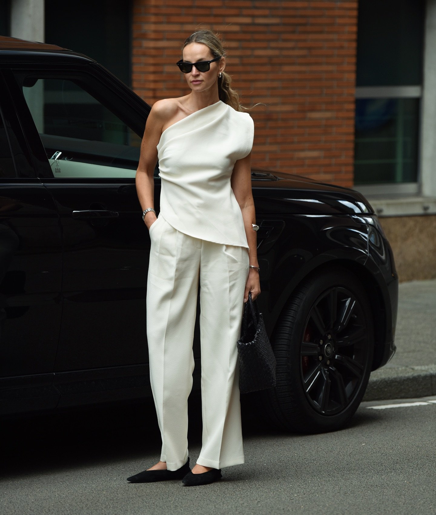 Mulher em look sofisticado e minimalista, vestindo conjunto branco com top de um ombro só e calça reta. Acessórios incluem óculos escuros elegantes e bolsa preta texturizada. Estilo moderno e refinado.