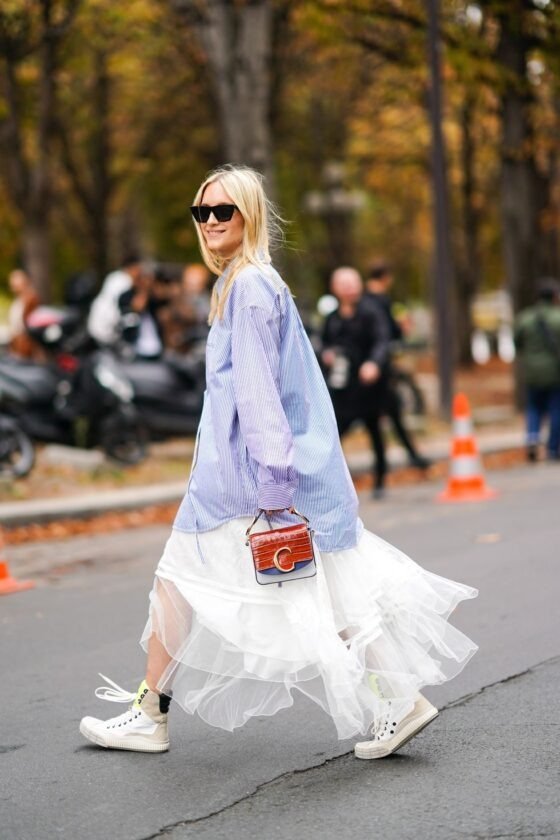 Estilo descontraído com sobreposição: camisa listrada oversized, saia de tule branco e tênis brancos. Acessório moderno com bolsa estruturada pequena. Tendência de equilíbrio entre o casual e o sofisticado.