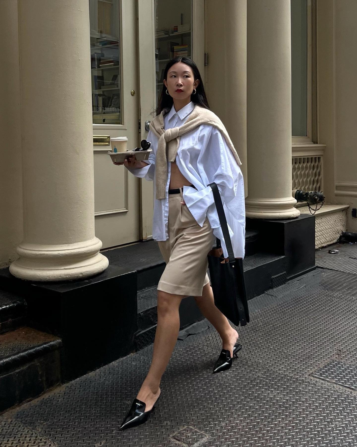 Mulher elegante caminhando com bermuda alfaiataria bege e camisa, carregando uma bandeja e bolsa preta.