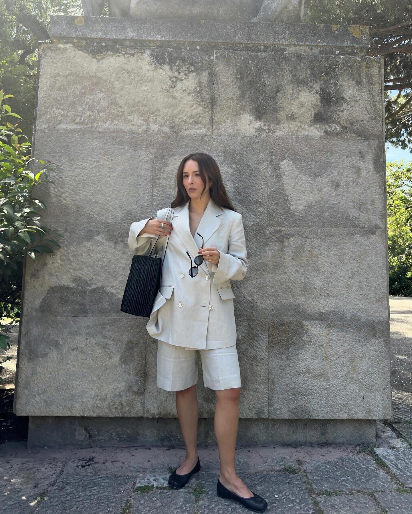 Pessoa usa conjunto de blazer e bermuda alfaiataria bege, segura bolsa preta e óculos, em frente a parede de pedra.