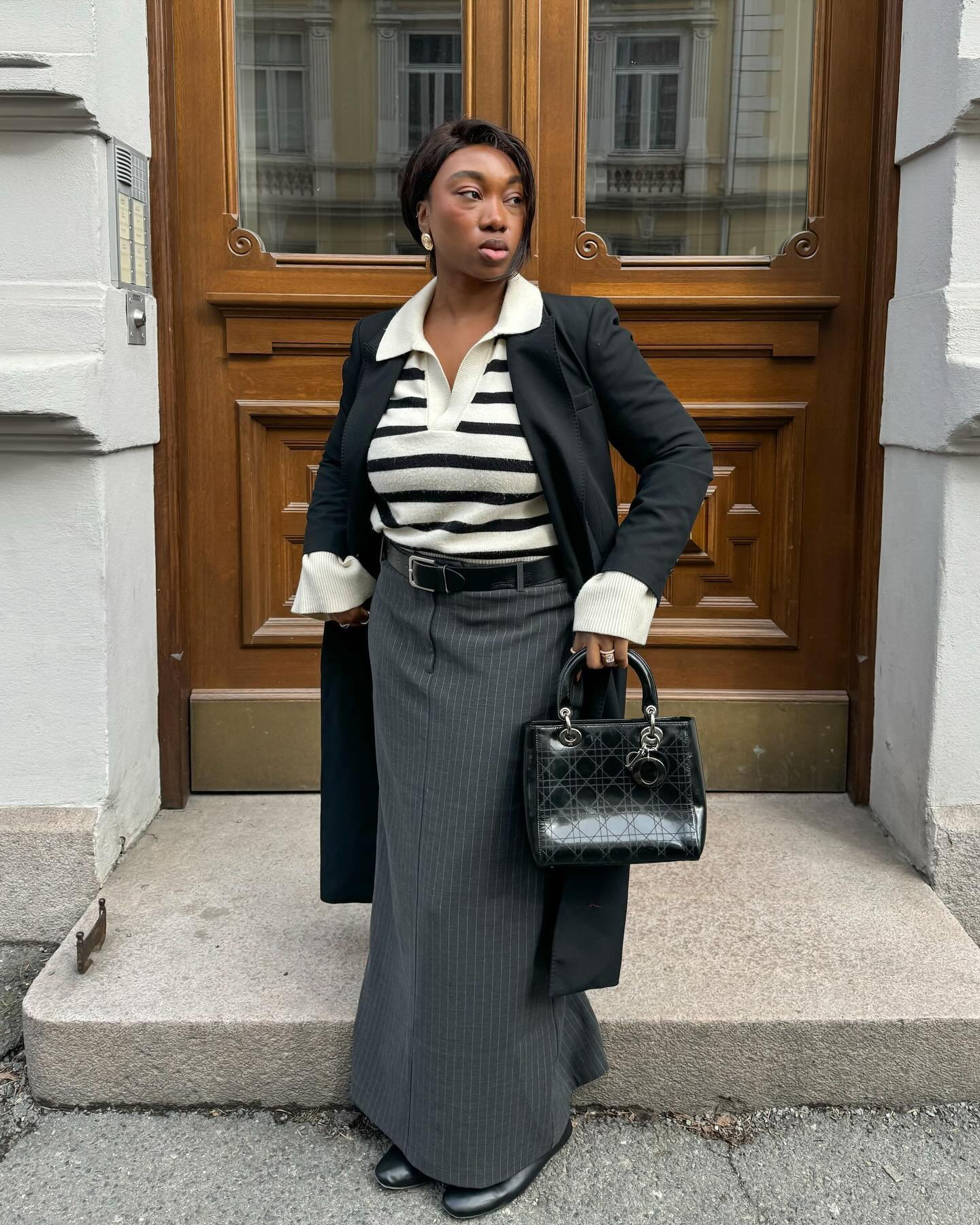 Mulher usando uma camisa polo listrada preta e branca, casaco preto, saia cinza e bolsa preta em frente a uma porta de madeira.