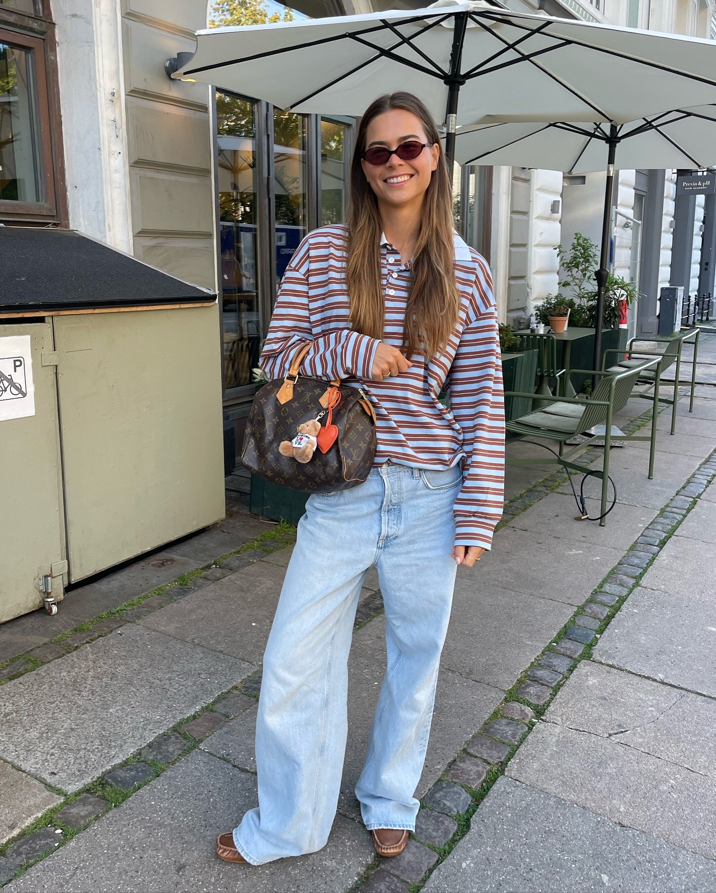 Pessoa sorrindo usando óculos de sol e camisa polo listrada, segurando uma bolsa. Fundo: ambiente urbano com mesas externas.