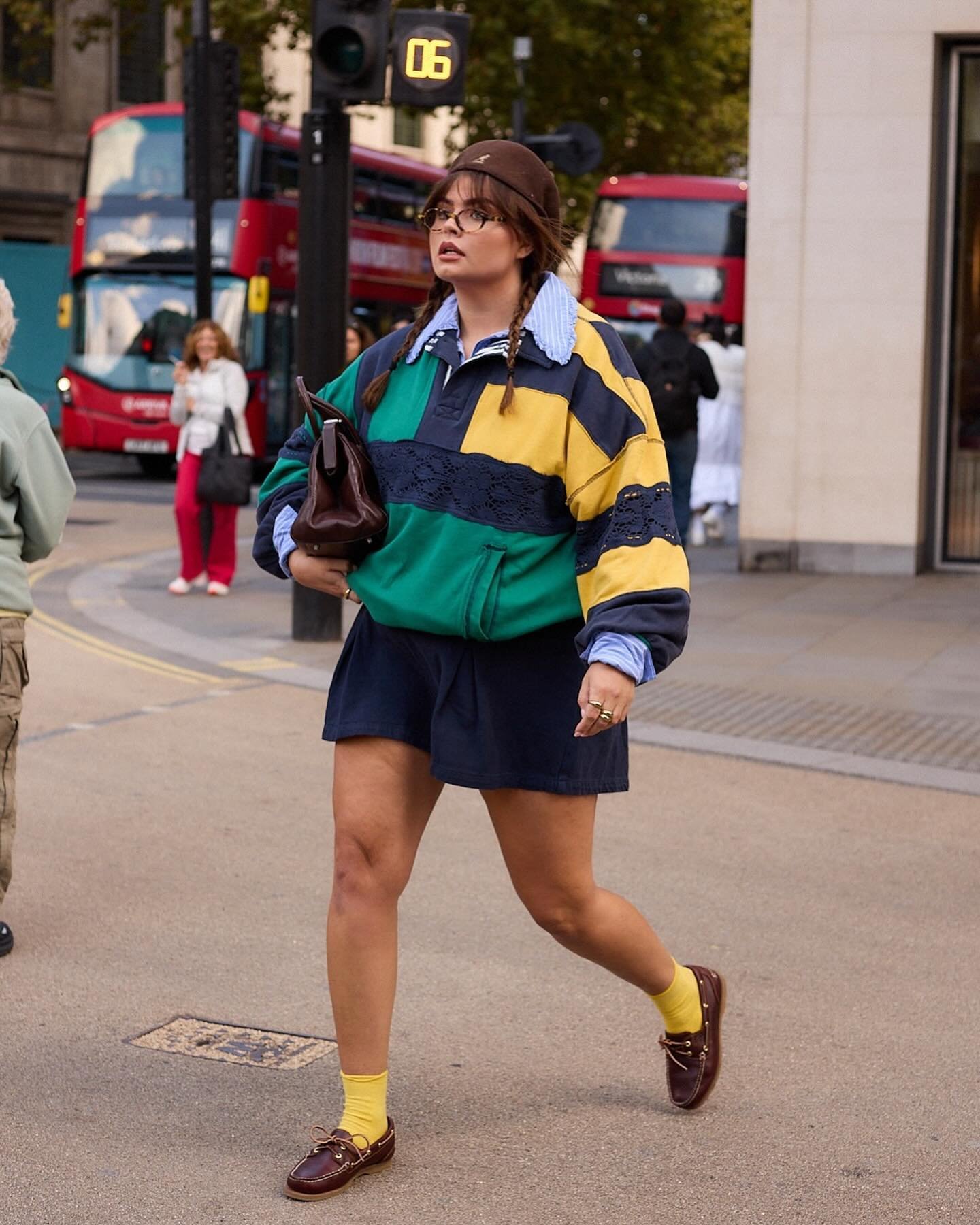 Pessoa caminhando pela rua em Londres, usando uma jaqueta colorida listrada sobre camisa polo, saia escura e meias amarelas.