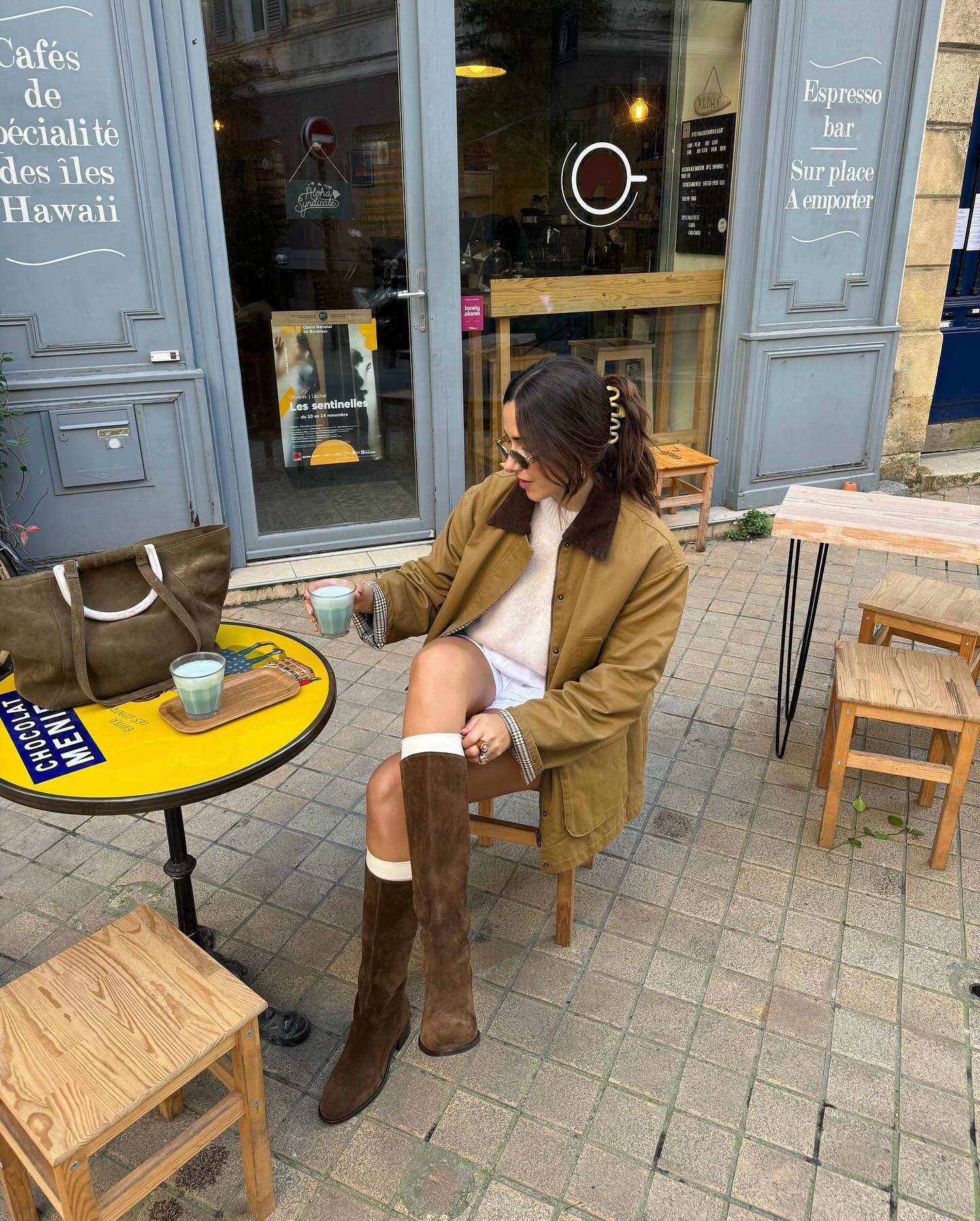 Mulher em um café usa botas de camurça marrons e casaco bege, com bolsa combinando ao lado.