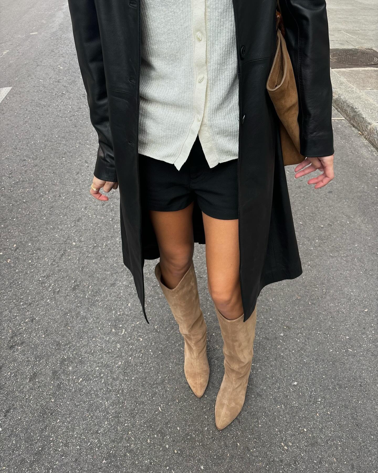 Pessoa usando botas de camurça bege, casaco longo preto, camisa branca e shorts pretos na rua.