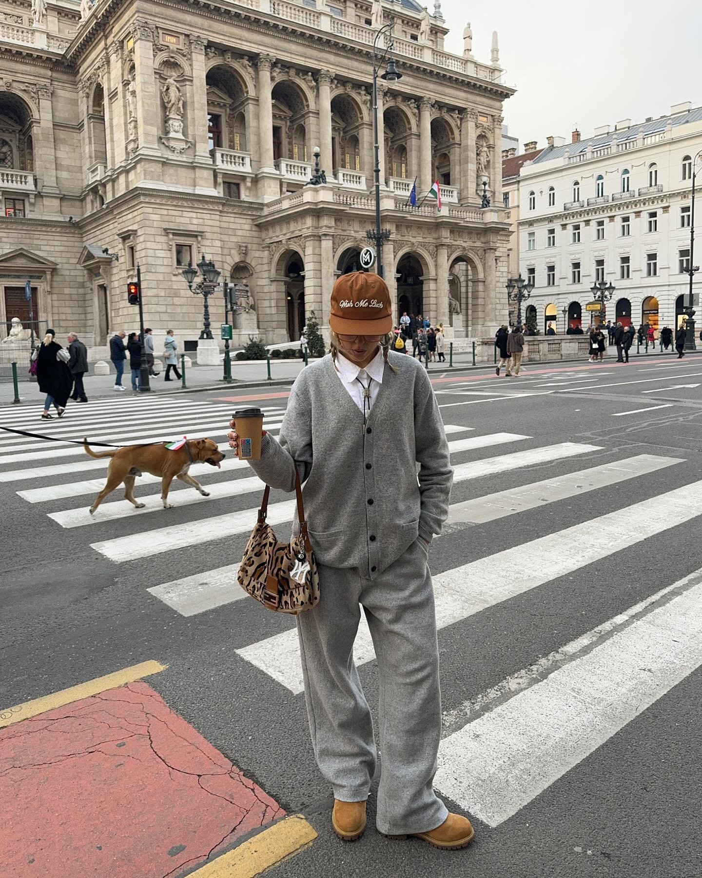 Pessoa vestindo moletom cinza passeia em frente a prédio histórico, segurando café e usando boné.