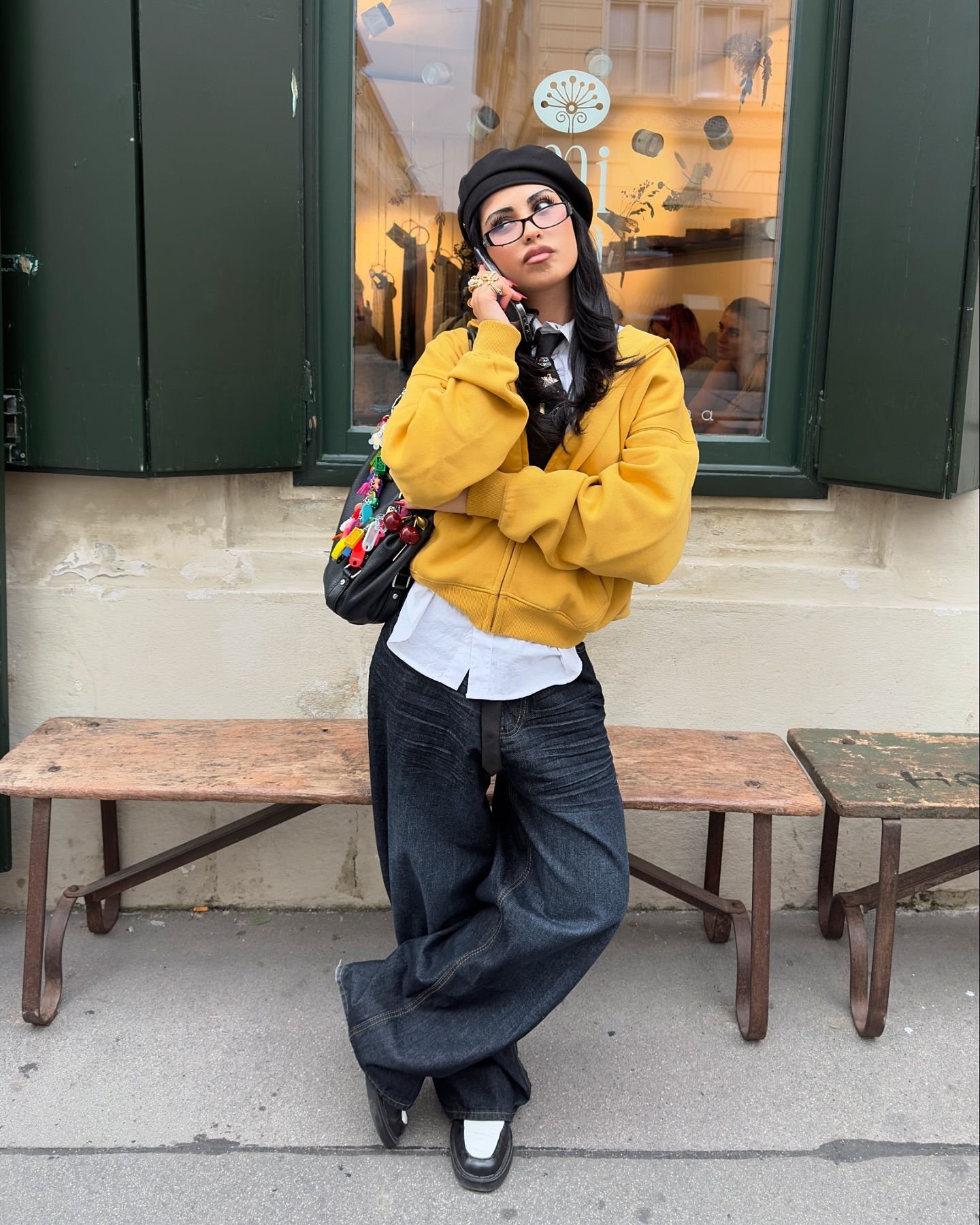 Mulher com moletom amarelo, calça jeans larga e boina preta. Mochila com chaveiros coloridos.