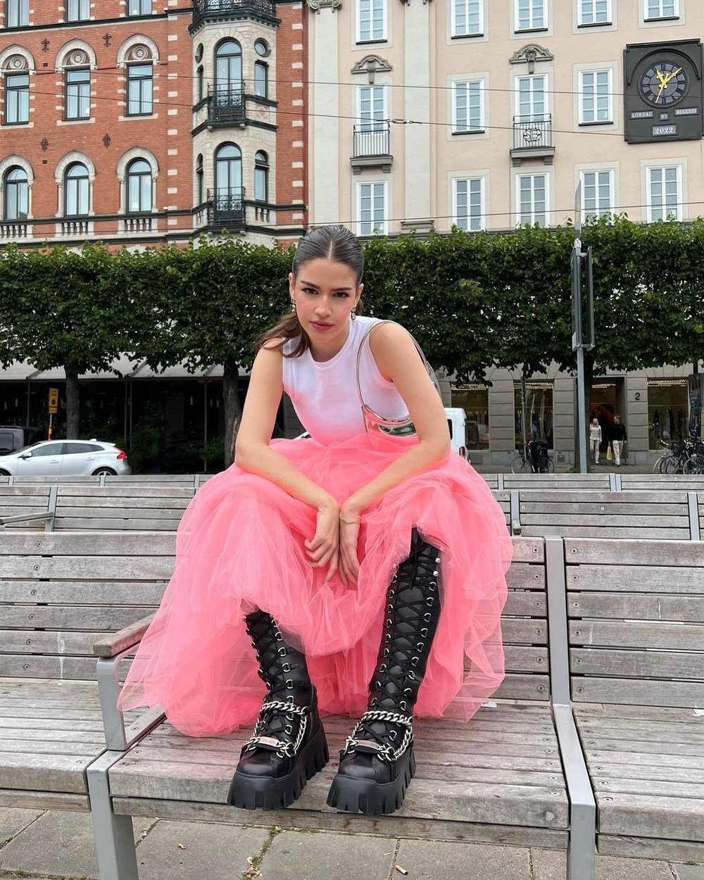 Pessoa vestindo saia de tule rosa, blusa branca e botas pretas de plataforma com correntes, sentado em banco ao ar livre. Visual mistura tendências punk e romântica, evidenciando contraste entre leveza e rebeldia urbana.