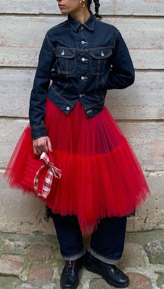 Pessoa com jaqueta jeans, saia de tule vermelho e calça preta. Bolsa vermelha com laço xadrez complementa o visual. Combinação criativa entre o urbano e o romântico, destacando inovação nas tendências de moda.