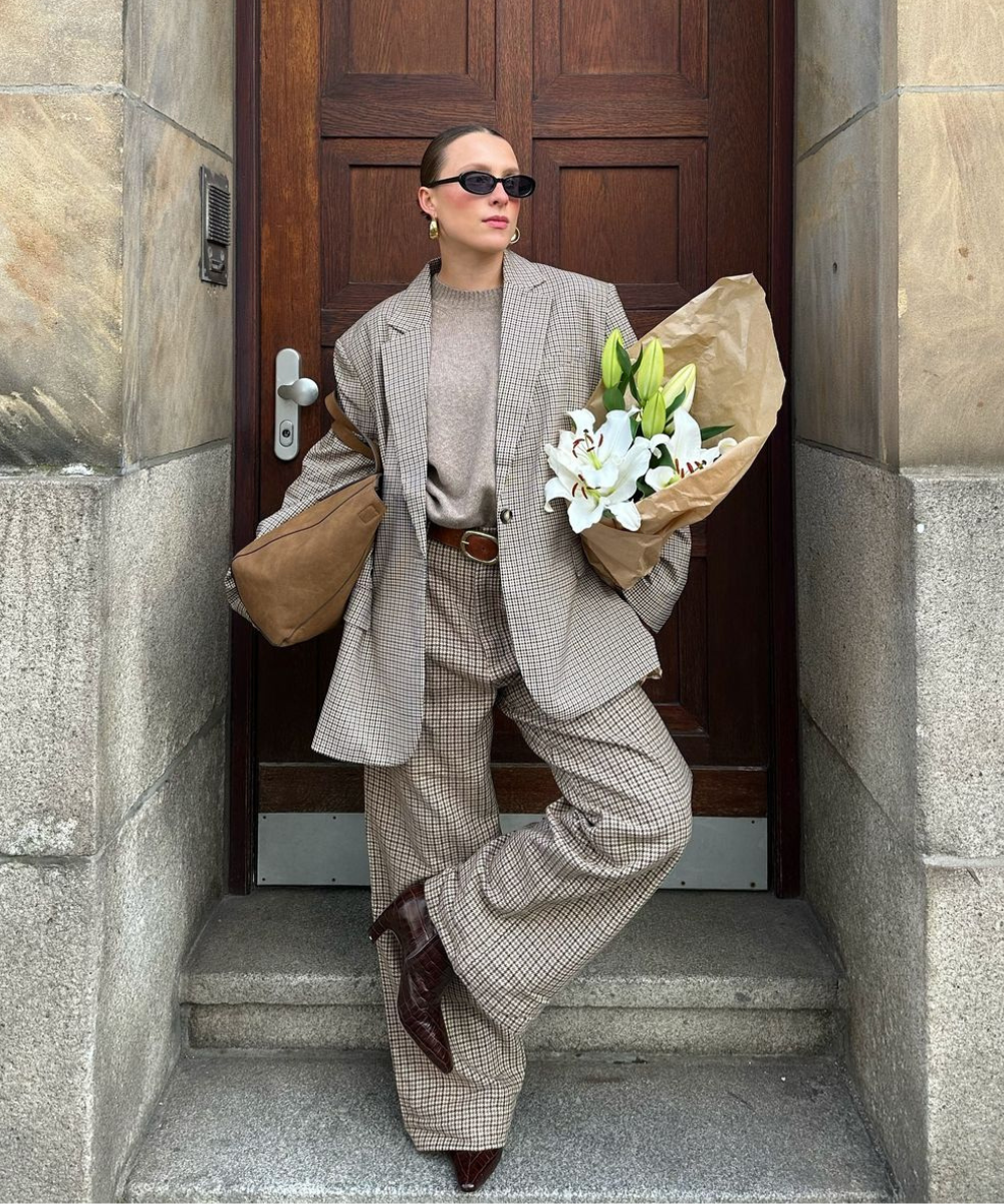 Pessoa com um dos looks com terninho da pauta. Segura buquê de flores na frente de porta de madeira.