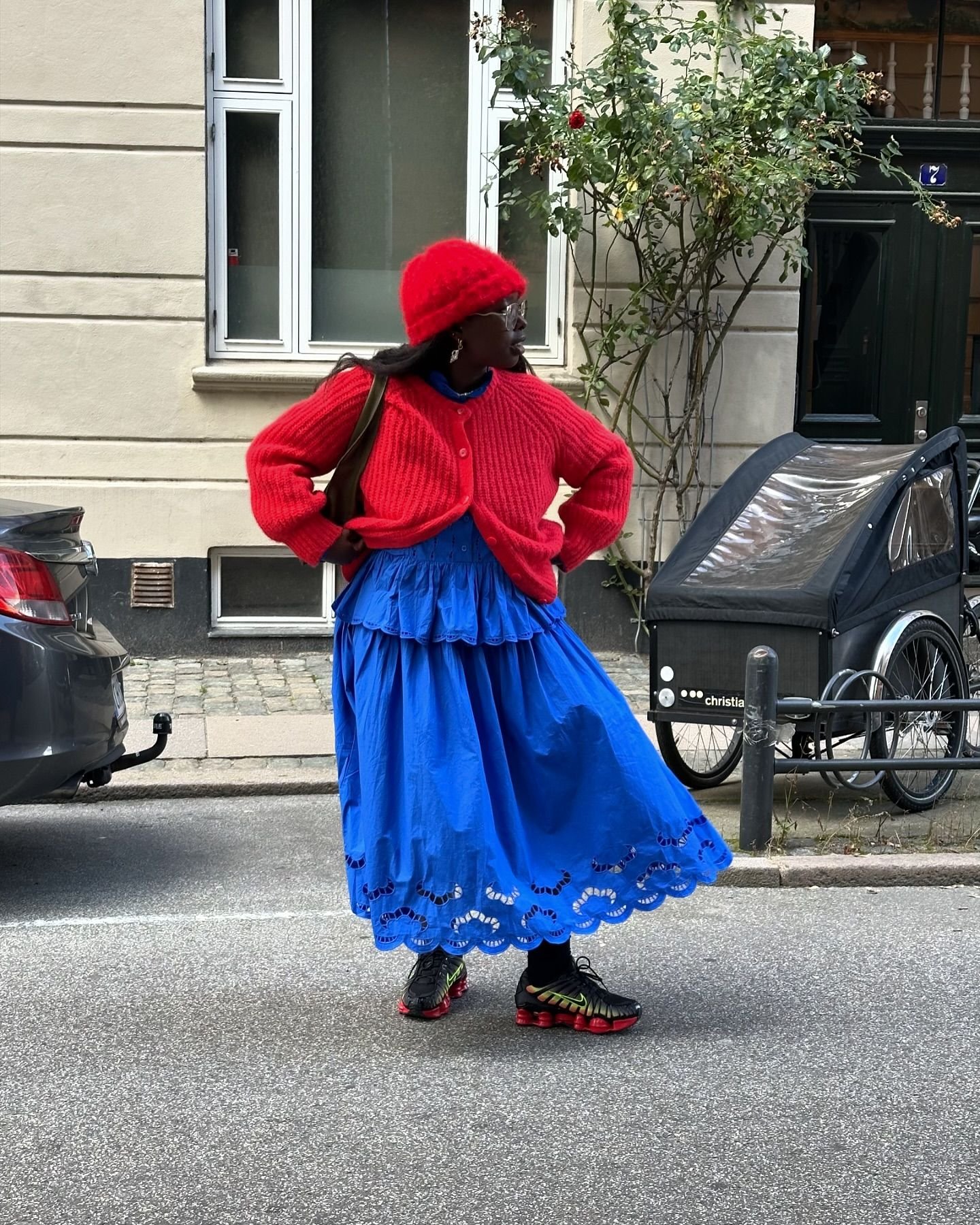 Pessoa em look colorido com saia azul e casaco vermelho, usando tênis Nike Shox, posa em rua urbana.