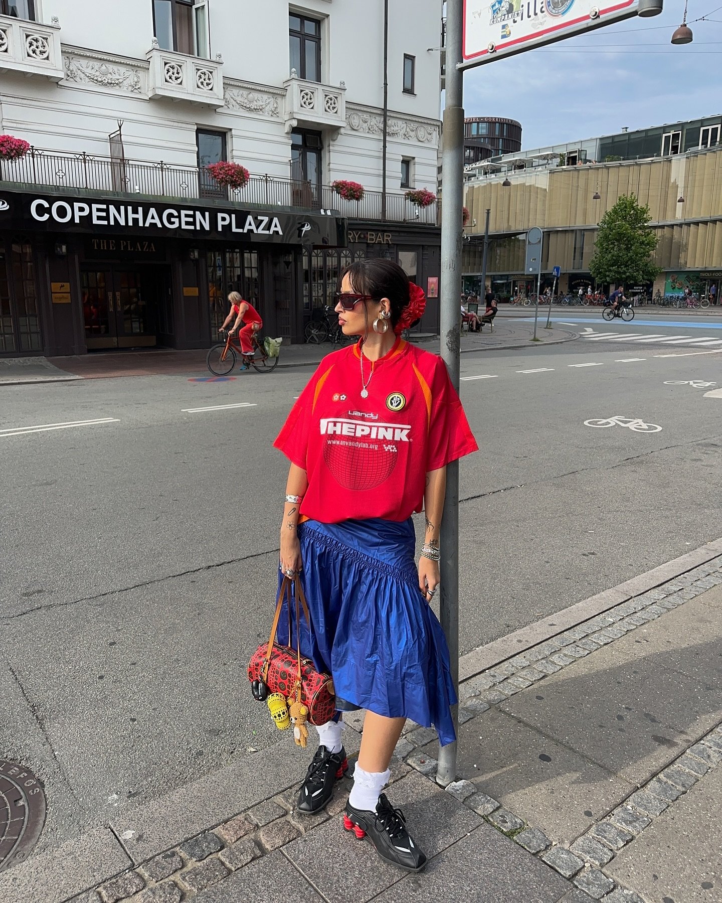 Pessoa com camiseta vermelha, saia azul e tênis Nike Shox em frente ao Copenhagen Plaza.