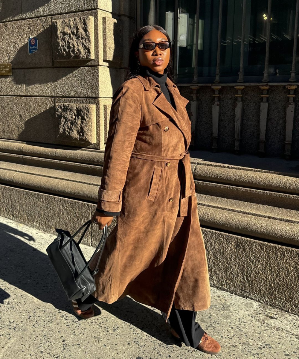 Pessoa usando trench coat marrom de camurça, com óculos escuros e bolsa preta. O look moderno e sofisticado destaca tendências de moda urbana, combinando elegância clássica com acessórios contemporâneos.