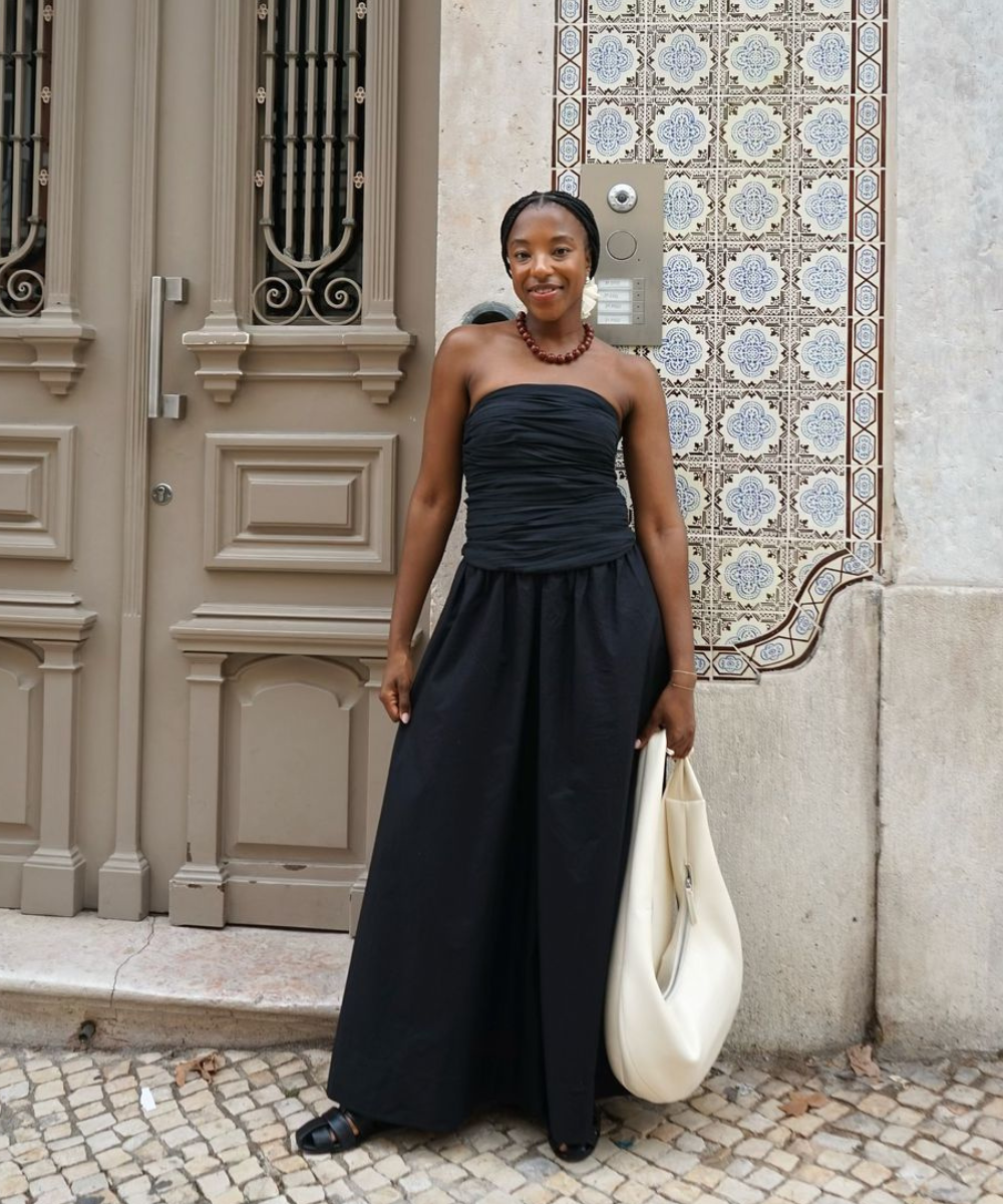 Pessoa sorridente em frente a uma porta e azulejos, vestindo look preto estiloso e segurando bolsa clara.