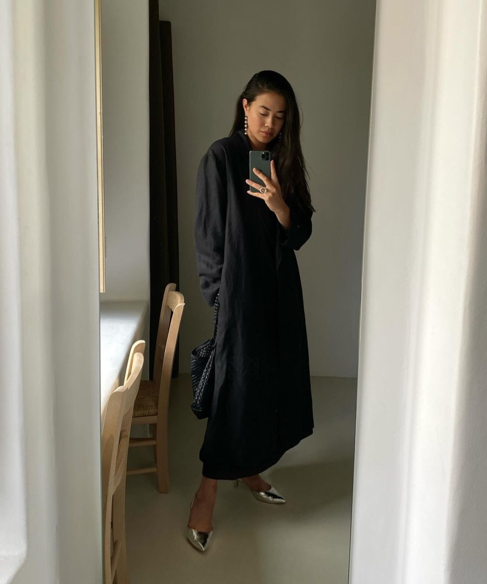 Looks pretos estilosos: Pessoa em traje preto estiloso, com casaco longo e sapatos prateados, tirando uma selfie em um ambiente minimalista.