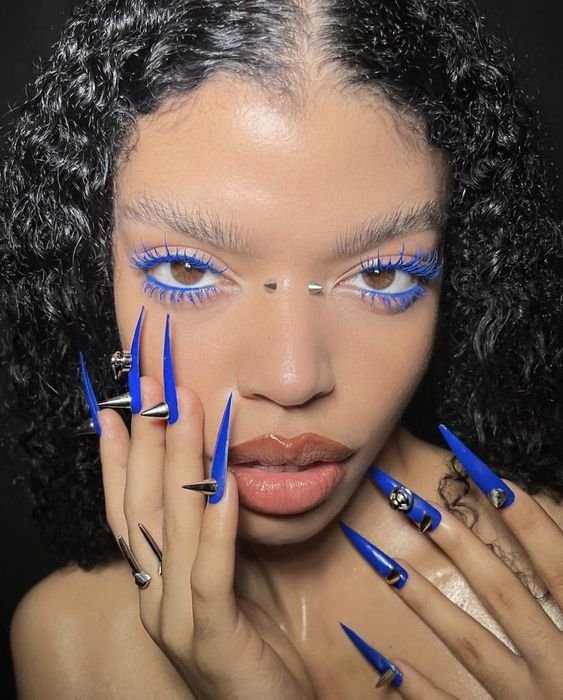 Unhas pontiagudas azuis e maquiagem impactante com cílios azuis e piercings são destaque. Estilo ousado e vanguardista, capturando tendências de moda contemporânea e expressão individual com acessórios marcantes.