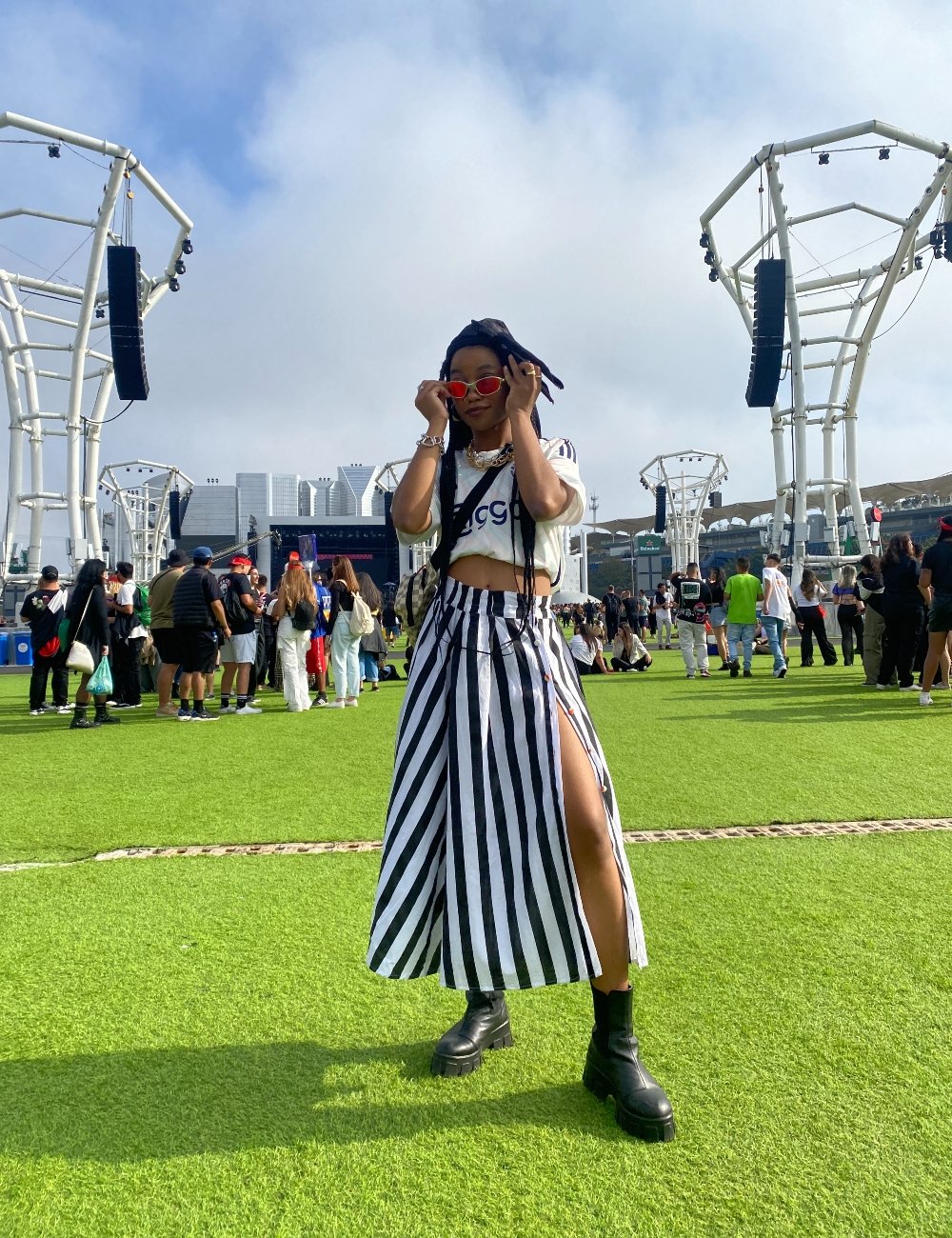 Mulher em festival ao ar livre usando top cropped branco, saia listrada preta e branca de corte reto com fenda lateral e botas pretas chunky. Acessórios incluem óculos de sol vermelhos e colares de metal. Favela