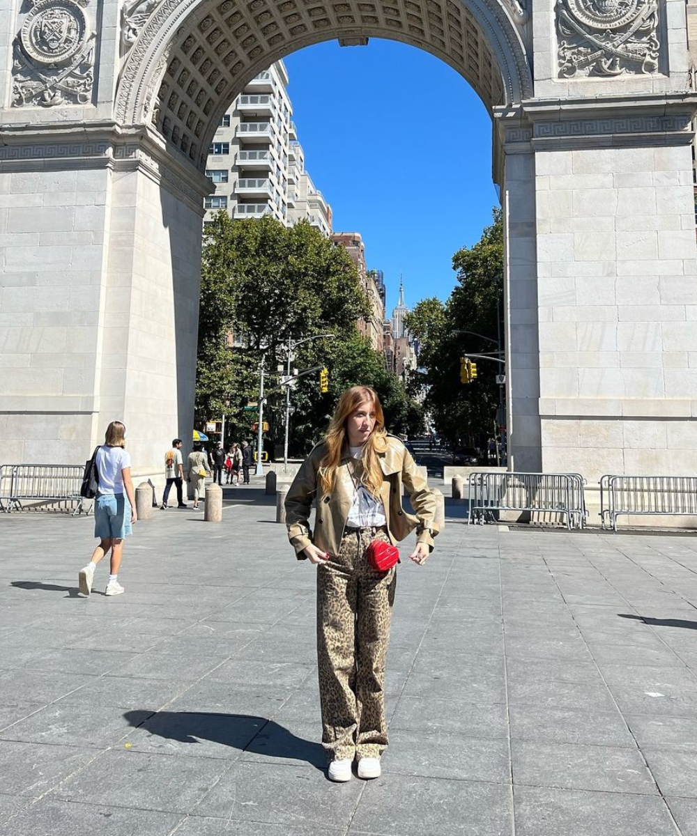 Pessoa de pé sob arco, com casaco bege e calça de estampado animal, carregando bolsa vermelha. Estilo pessoal vibrante.