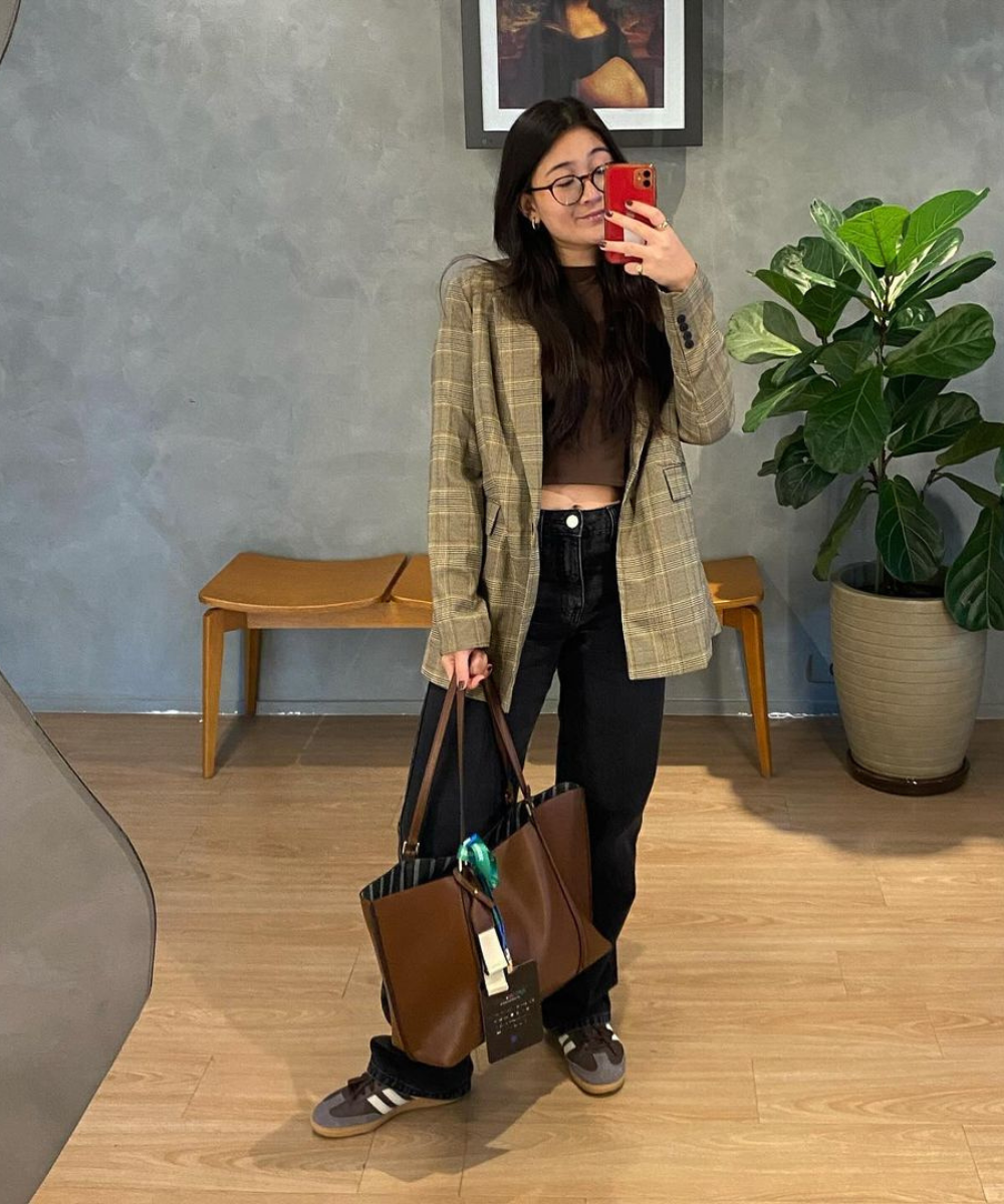 Jovem tirando selfie com estilo casual: blazer xadrez, calça preta, bolsa grande. Estilo pessoal evidente na escolha do look.