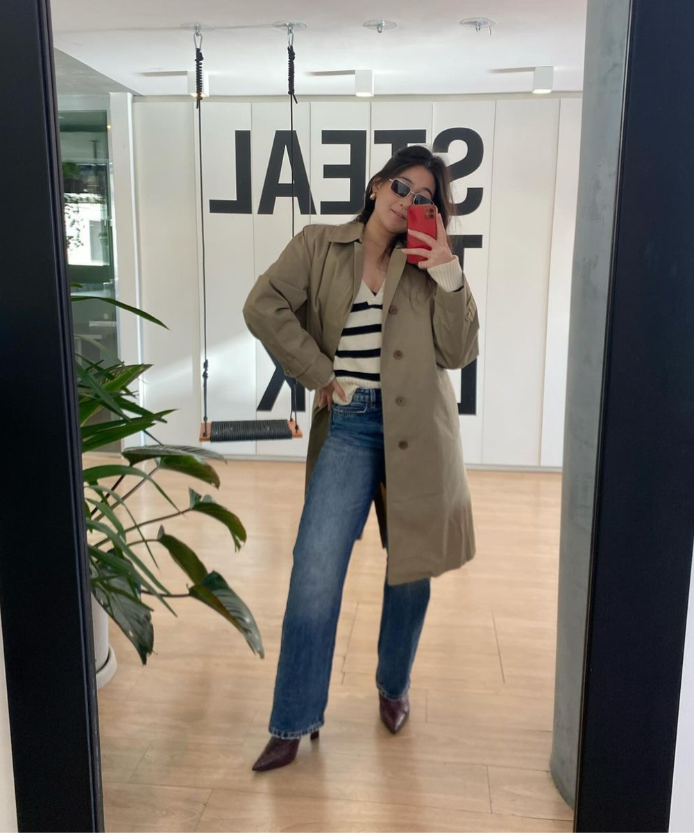 Pessoa tira selfie em frente ao espelho usando trench coat, calça jeans e blusa listrada, reflete estilo pessoal.