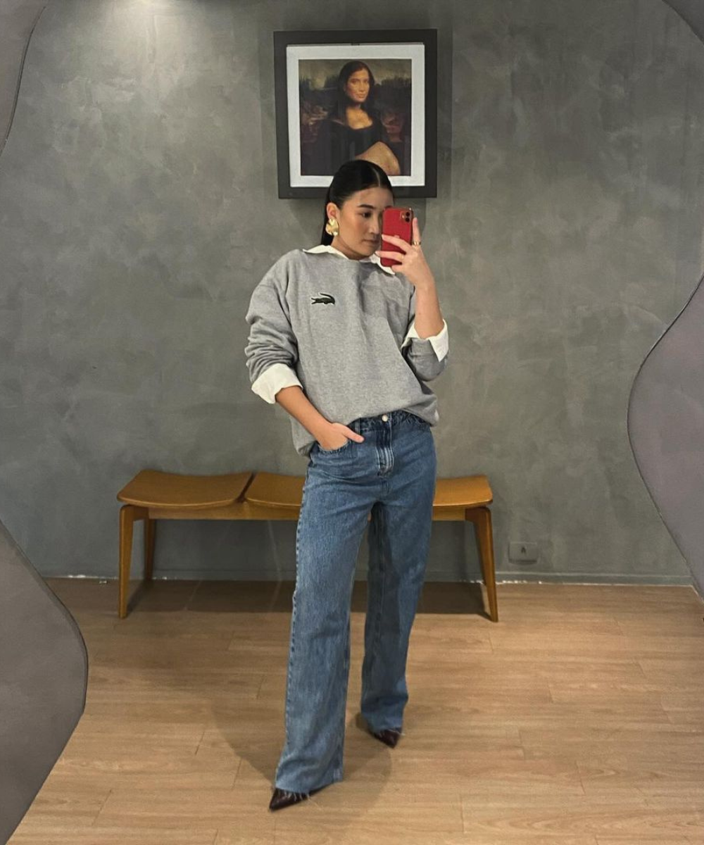 Pessoa usando moletom cinza e jeans, tirando selfie em frente a espelho. Estilo pessoal casual, com quadro ao fundo.