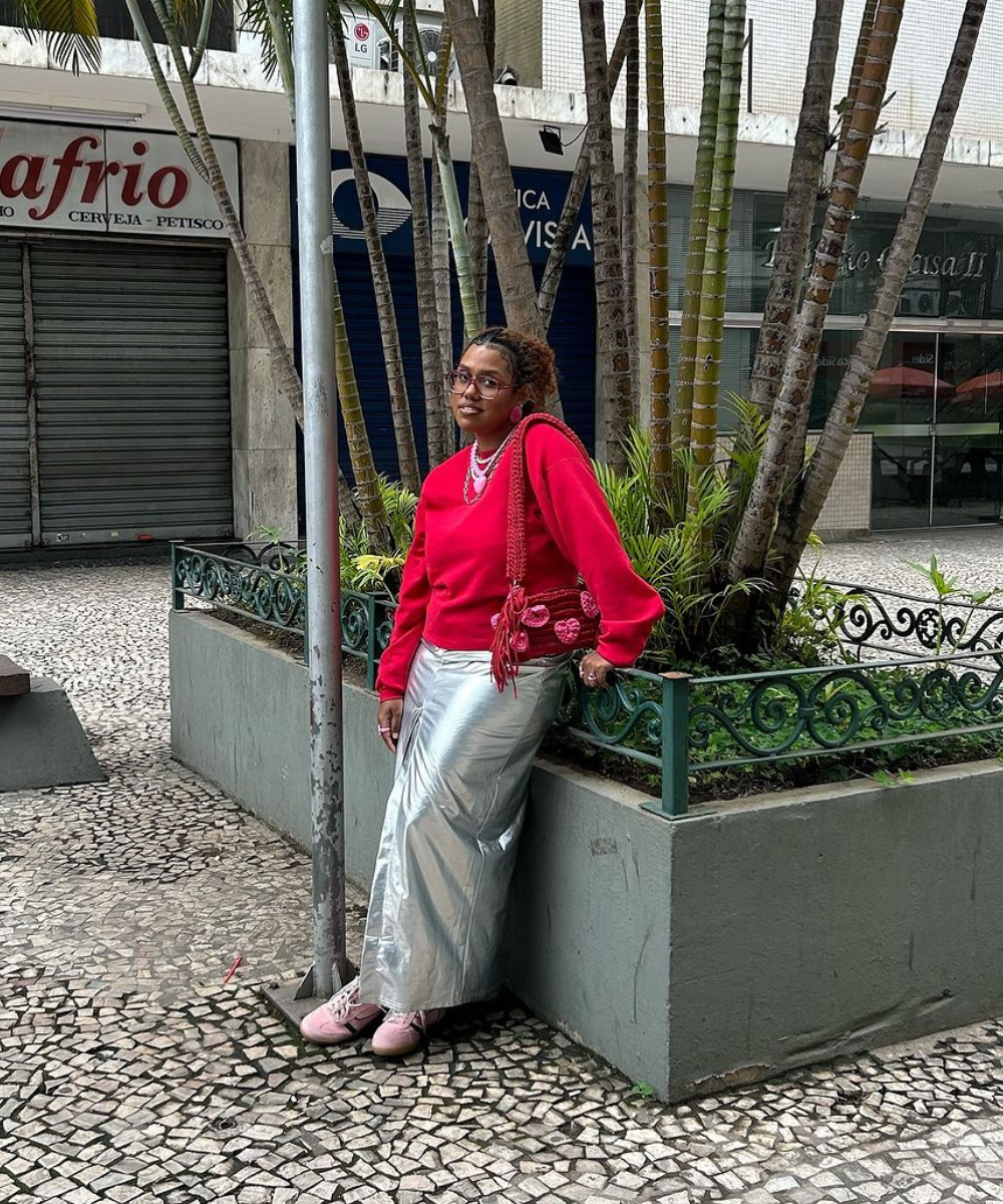 Uma pessoa usando blusa vermelha e saia prateada posando em frente a uma plantação urbana, destacando estilo pessoal.