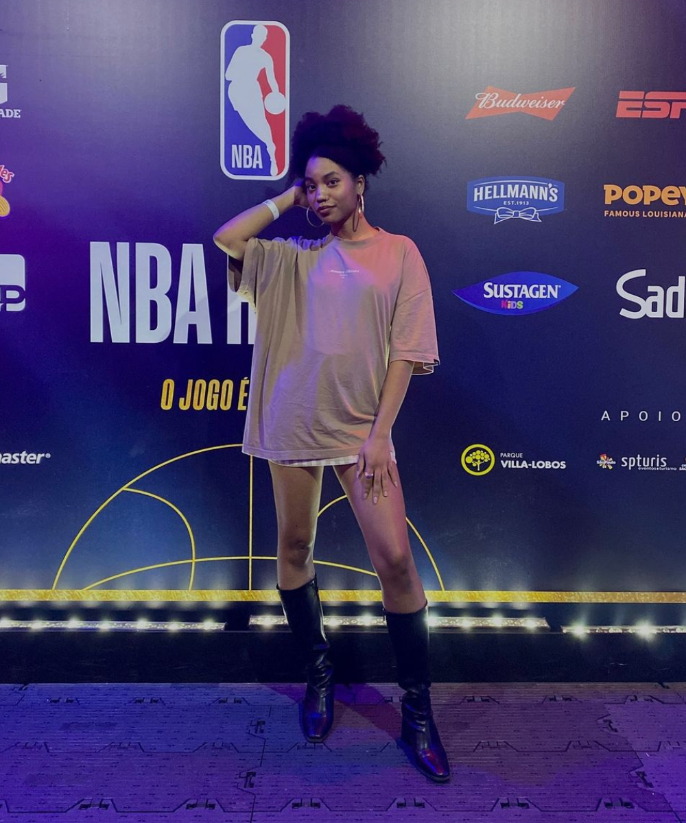 Pessoa posando em evento da NBA, vestindo camiseta larga e botas, destacando seu estilo pessoal único. Banners de marcas no fundo.