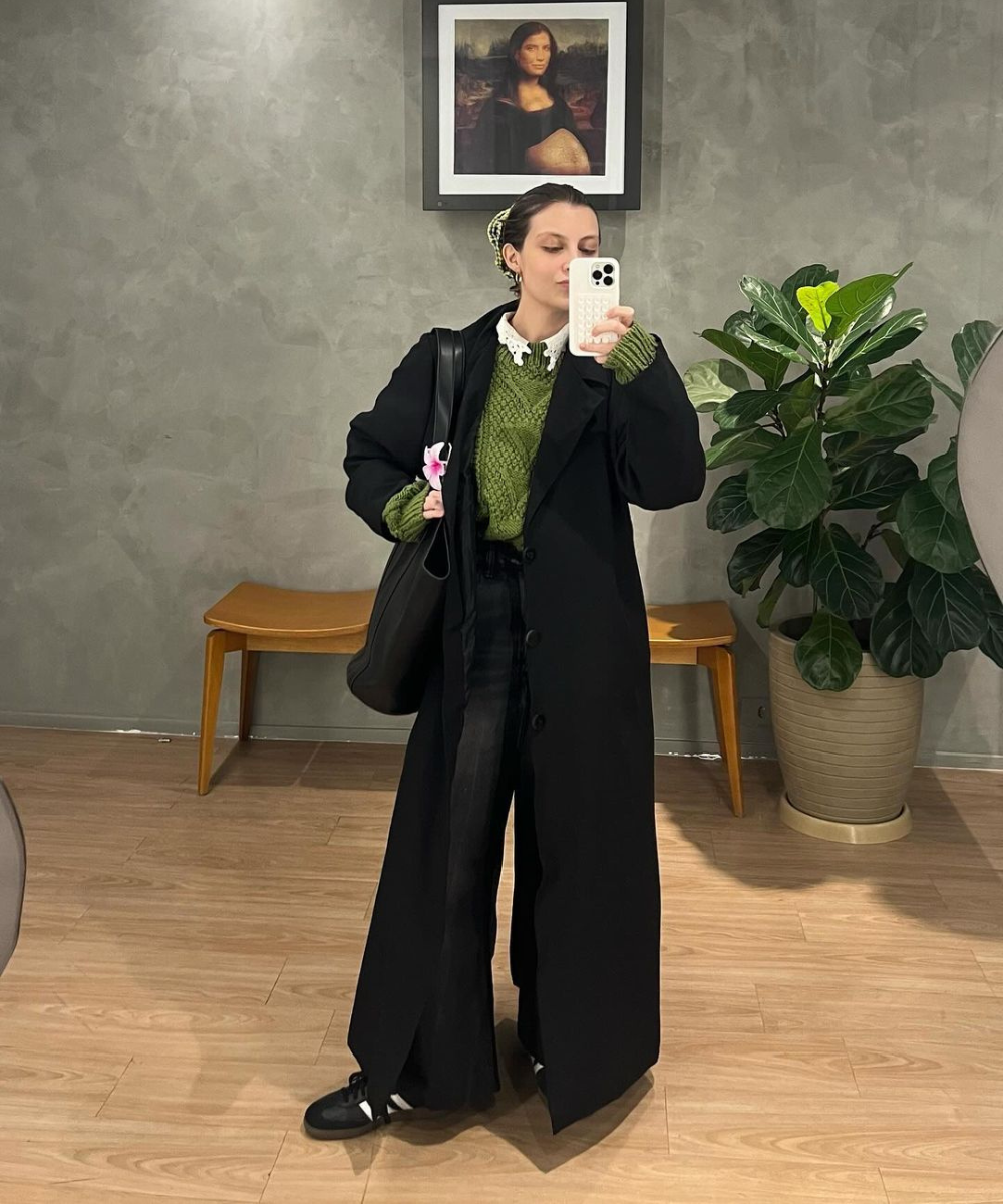 Pessoa com casaco preto longo e suéter verde tirando selfie num hall. Estilo pessoal moderno.