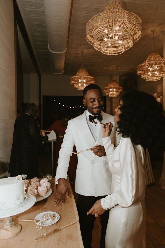 Casal elegantemente vestido: ele de smoking branco com gravata borboleta, ela com vestido de cetim longo e elegante. Estilo clássico e atemporal, destacando-se em um ambiente sofisticado com lustres de palha rústica. Mini wedding.