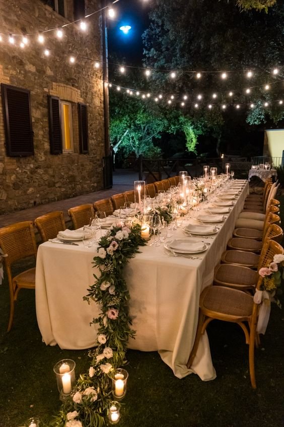 Mesa de jantar ao ar livre em estilo rústico-chique, com toalha branca, arranjo de flores e velas, sob luzes pendentes. Tendência em decoração de casamentos e eventos ao ar livre. Mini wedding.