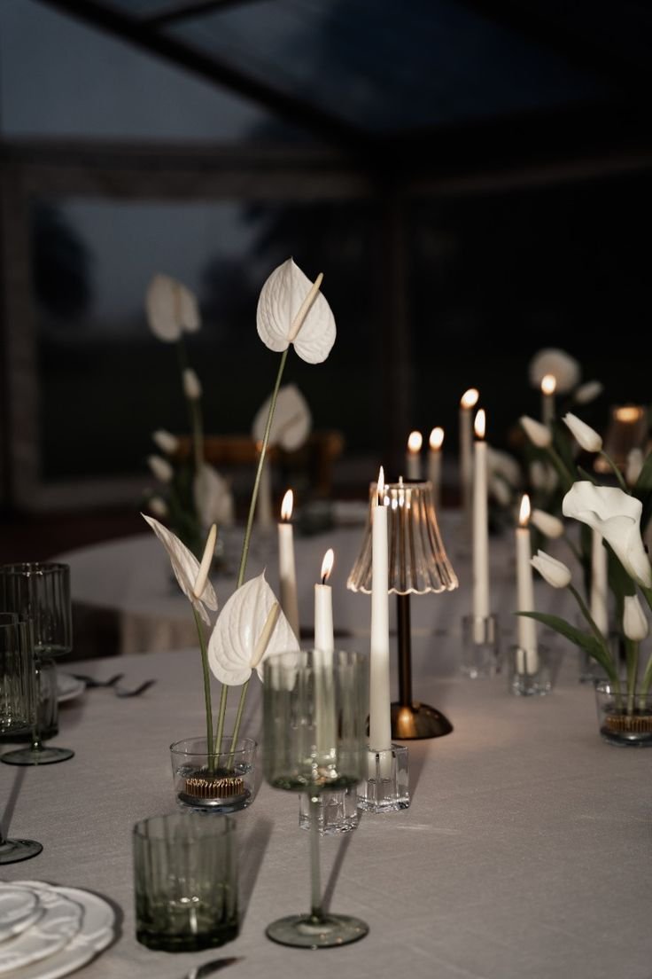 Mesa de jantar elegante com copos de vidro esverdeados, velas brancas altas e arranjos florais minimalistas. Tendência sofisticada em decoração de interiores, destacando o uso de tons neutros e iluminação suave. Mini Wedding