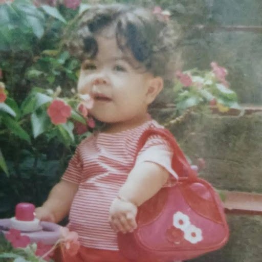 Criança sorrindo em jardim florido, usando roupa vermelha listrada e segurando uma bolsa infantil.