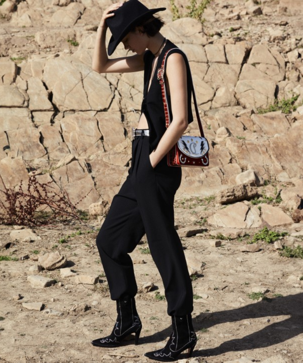 Pessoa usando traje preto e chapéu, caminhando em terreno rochoso. Bolsa vermelha com detalhes. Novidades de Outubro.