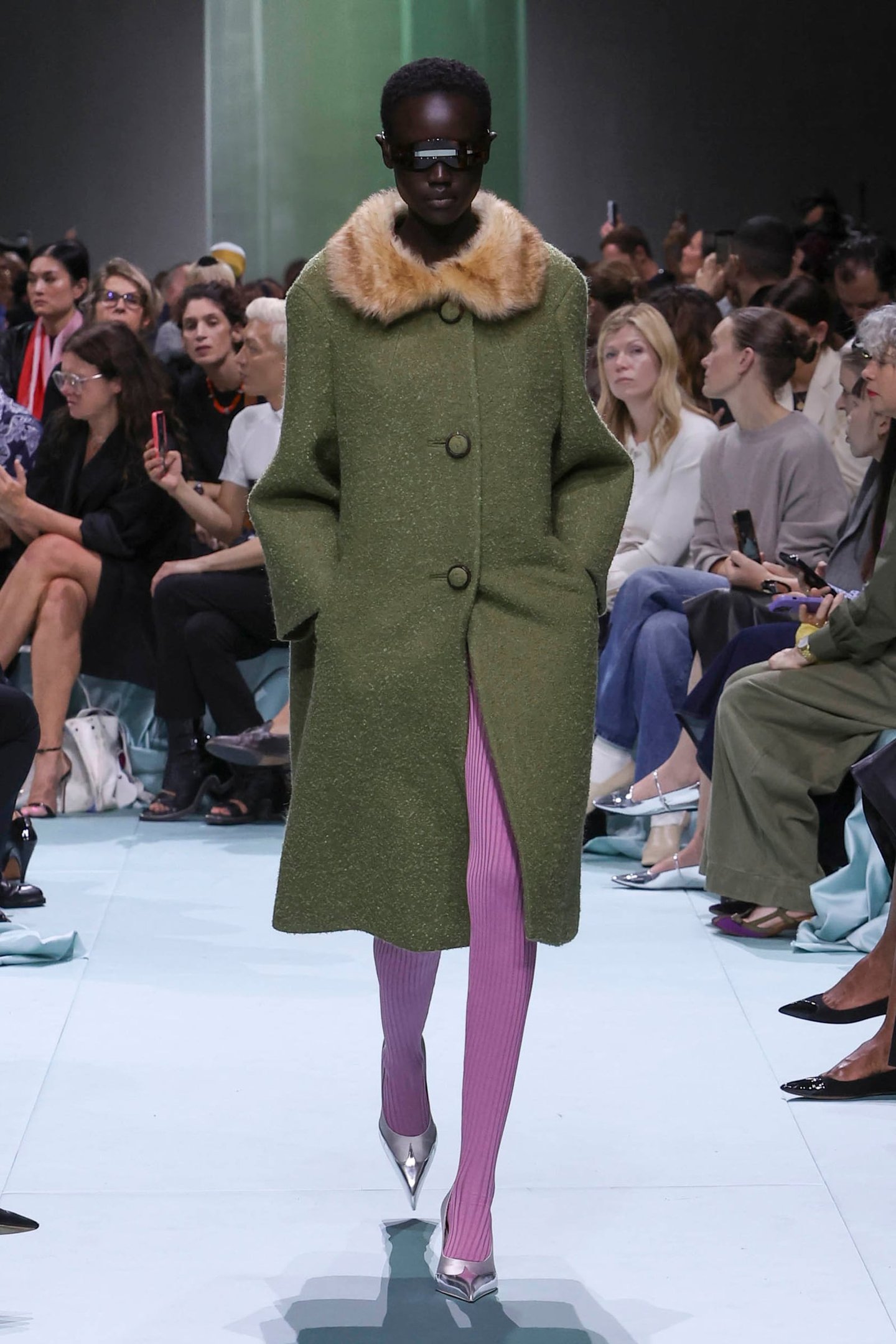 Desfile de moda com casaco verde de tecido pesado e gola de pele sintética, combinado com meia-calça rosa e sapatos prateados. Tendências incluem contrastes de textura e cores ousadas, destacando acessórios modernos como óculos futuristas.