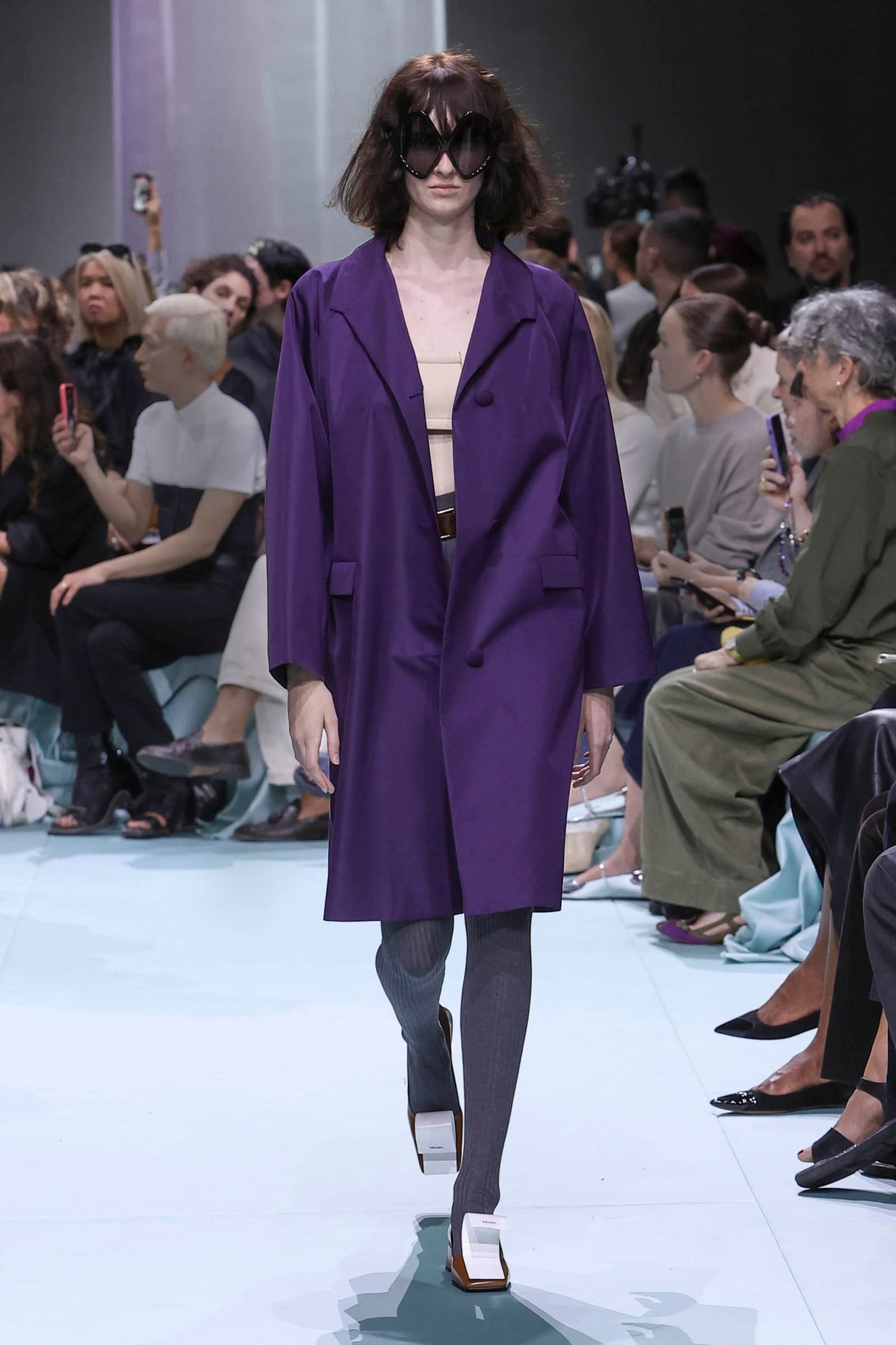 Modelo veste blazer roxo oversized sobre top nude, meias cinza longas e sapatos de design geométrico branco. Óculos escuros grandes adicionam um toque dramático, destacando tendências de moda como o maximalismo e o minimalismo contemporâneo.