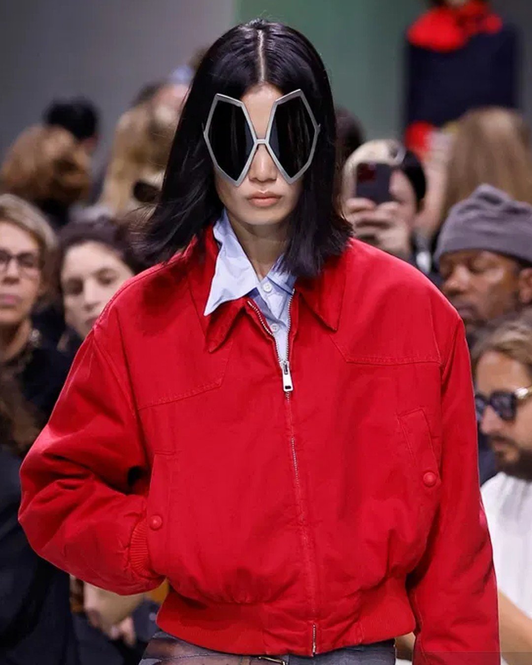 Desfile de moda com foco em tendências ousadas: modelo veste jaqueta vermelha oversized e camisa azul clara, combinando com óculos de sol de design futurista e amplo destaque. Look moderno e impactante.