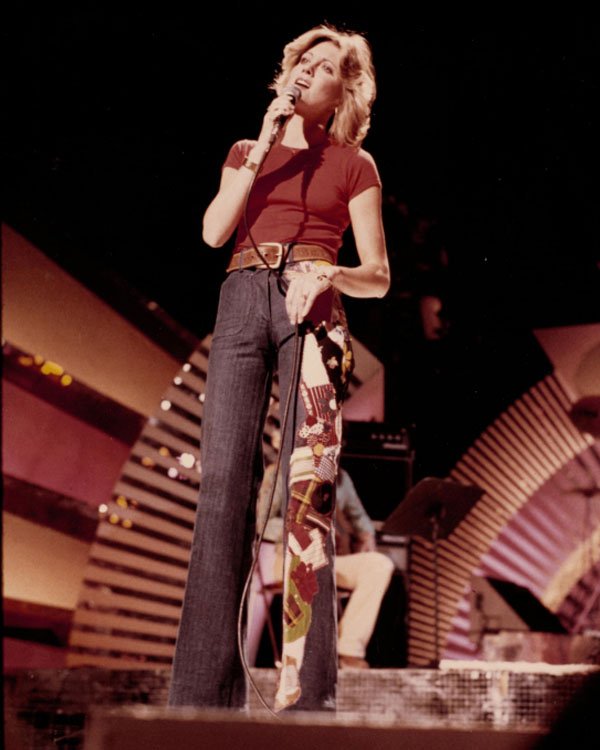 Cantora no palco com jeans boca de sino e blusa vermelha, em estilo clássico dos anos 70.